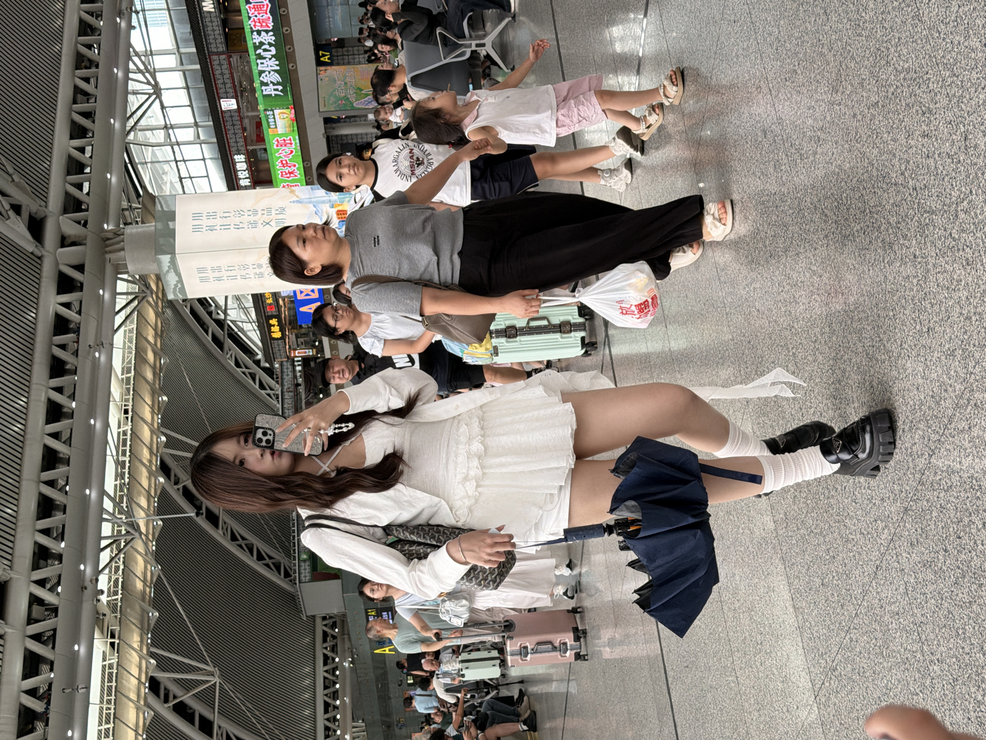 In Panyu District, China, within a spacious, modern indoor transit terminal with a high, arched ceiling structure and bright lighting, a young woman with long, dark hair is walking and taking a selfie. She is wearing a white ruffled dress and a white jacket, white socks, and black platform loafers. In her left hand, she holds a dark umbrella that is folded and secured with a ribbon, and in her right hand, she holds her smartphone. She is surrounded by other travelers. To her right, a woman in a gray t-shirt and black pants is walking, carrying a white plastic bag. Behind them, a teenage girl in a white t-shirt and dark shorts is walking with a younger girl in a pink sleeveless top and pink shorts, holding the younger girl's hand. Other individuals and luggage are visible in the background, suggesting a busy travel hub.