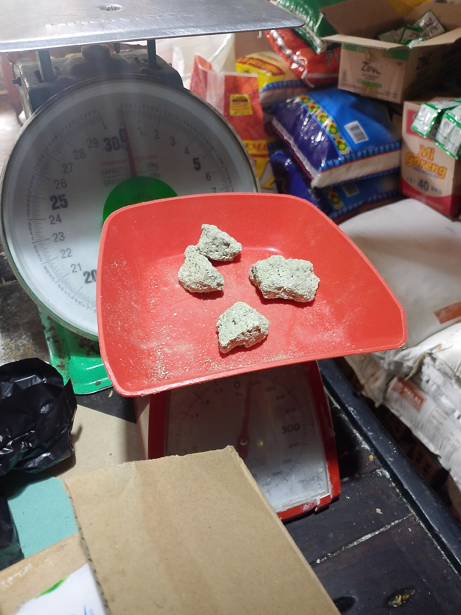 A close-up, eye-level shot captures a portion of a food stall in Palembang, Indonesia. In the foreground, a red plastic tray sits atop a green and red analog scale, displaying four irregular lumps of what appears to be dried material, possibly food ingredients or flavoring. The scale's face shows a reading slightly above 30, with markings up to 50 and a green indicator. The background is cluttered with various bags of food products, including bags of rice and boxes of instant noodles with Indonesian labels like "Mi Goreng." The overall environment suggests a bustling market or food preparation area, with visible dust and debris on the wooden surface below the scale.