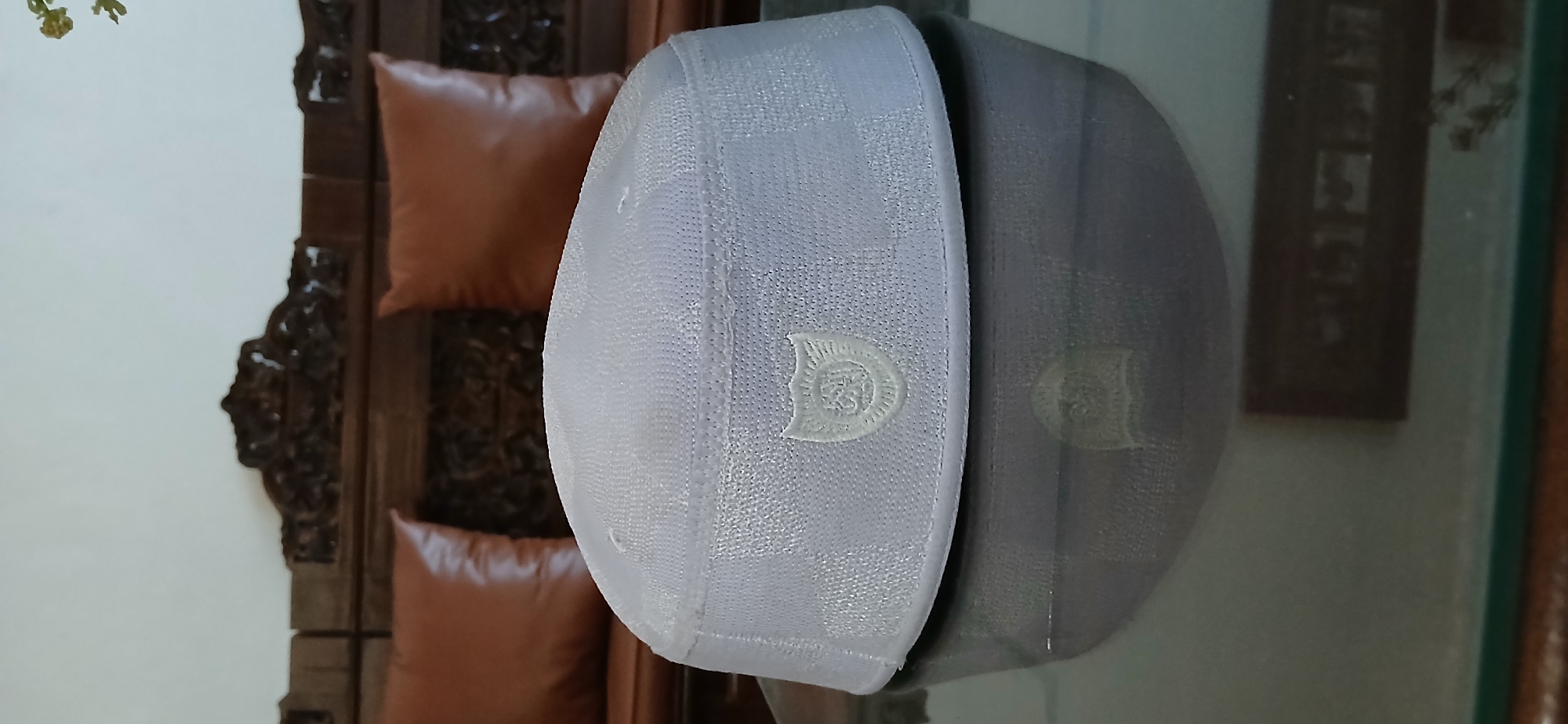 A white, woven skullcap is placed on a reflective surface, likely a table, in front of a wooden bench with brown cushions. The skullcap has an embroidered emblem on the front. The background appears to be an indoor setting, possibly a home, with a wall and part of a couch visible. There is no indication of the specific location within Samarinda, Indonesia.