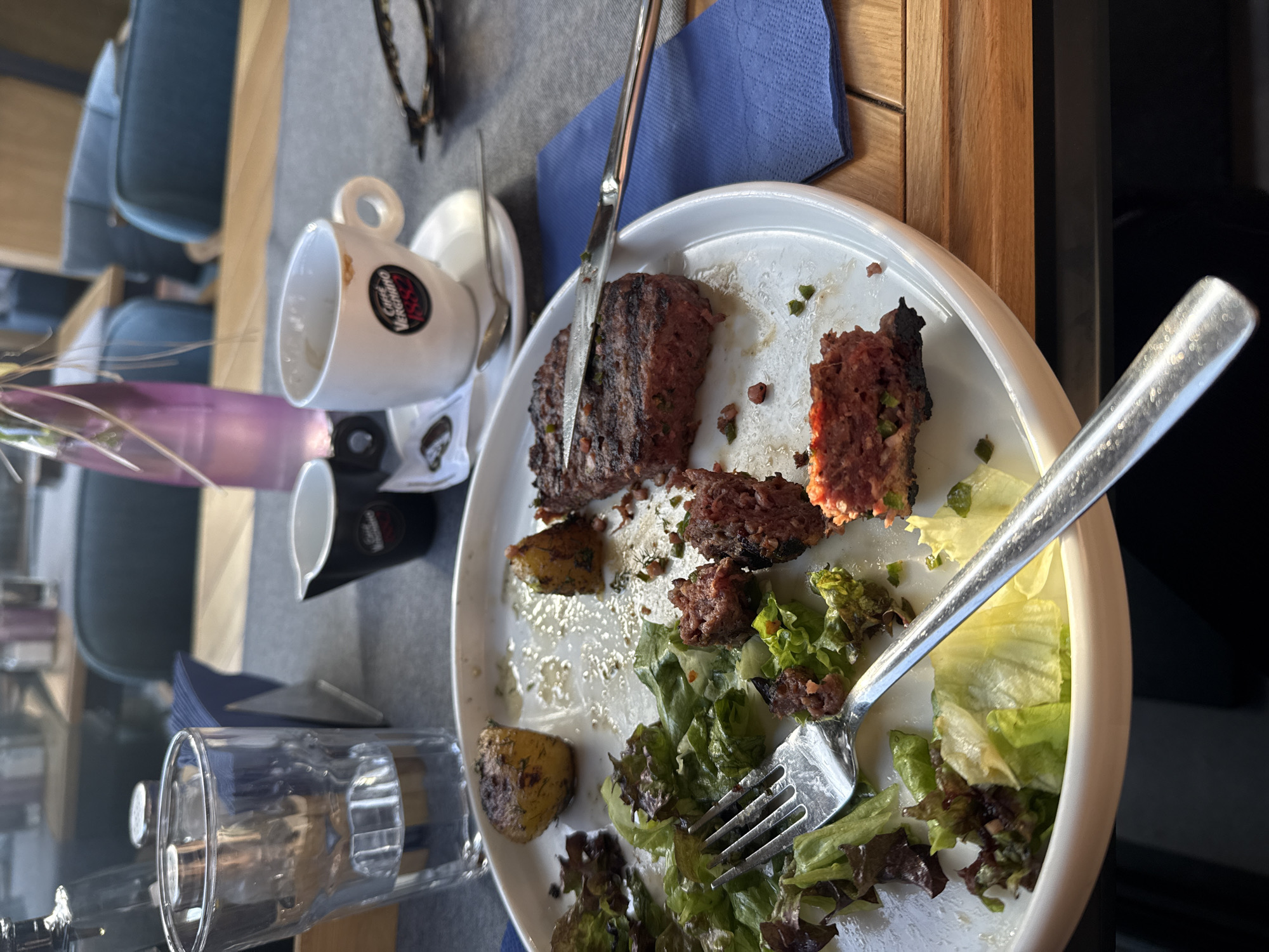 The image depicts an unfinished meal on a white plate, likely in a restaurant setting. The plate contains remnants of meatloaf, potatoes, and salad. A fork and knife rest on the plate, suggesting the meal was interrupted. Other items on the table include a coffee cup, creamer, glass, glasses, and a blue napkin. The setting appears to be in a cafe or restaurant based on the table setting and presence of beverages and food remnants. The exact location in Sofia, Bulgaria, cannot be determined from the image alone, as there are no distinctive landmarks.
