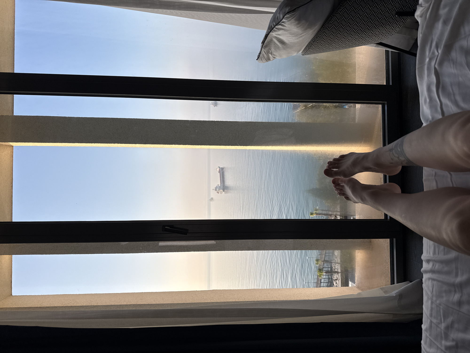 The image shows a first-person perspective of someone lying in bed, with their legs and feet visible in the foreground. They are looking out of a large window at a view of the sea with ships. The window has vertical dividers, creating multiple panes. The curtains are partially drawn on the left and right sides. The overall ambiance suggests a hotel room with a sea view. The location is identified as Limassol, Cyprus.