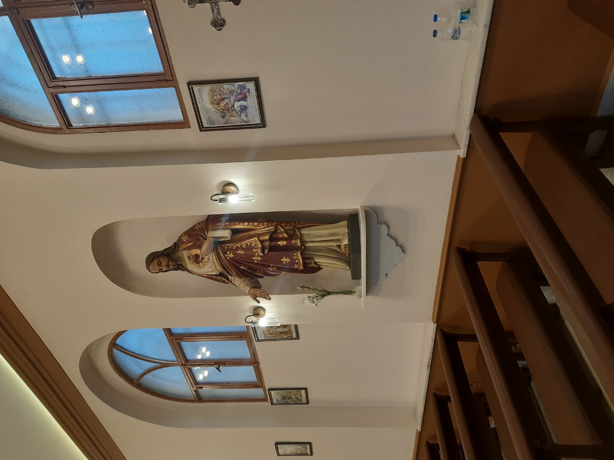 This image captures the serene interior of a religious building, likely a church or chapel, located in Kadıköy, Turkey. The central focus is a statue of Jesus Christ, depicted as the Sacred Heart, positioned within an arched alcove on a light-colored wall. Jesus is shown in maroon and gold robes, with his right hand extended forward and his left hand gesturing towards his exposed, flaming heart. A small vase of white baby's breath flowers rests on the ledge beneath the statue.
Above the statue, two modern-style wall sconces provide artificial light, while additional recessed lighting illuminates the ceiling. Two large, rectangular wooden-framed windows, filled with frosted or textured glass, are visible higher on the wall, allowing soft blue light to filter in from what appears to be a clear sky outside, suggesting daytime. Below these, another arched window with a wooden frame adds to the architectural detail.
Other religious elements include a decorative cross hanging from the ceiling in the upper right corner and several framed pictures on the walls. One prominent framed picture displays an artistic rendering of religious figures. Visible text on this picture identifies "MESİH İSA DÜNYANIN KURTARICISI" (Jesus Christ the Savior of the World), "THE SACRED HEART", "HAZRETİ MERYEM ANA, İSA'NIN ANNESİ" (Mother Mary, Mother of Jesus), and "VE O'NUN SEVGİLİ HAVARİSİ YUHANNA" (And His Beloved Disciple John).
To the right, a dark brown wooden railing or balustrade, possibly part of a staircase or upper floor, runs along the edge of the frame. On a white ledge above this railing, two plastic bottles of "Uludağ" brand mineral water are casually placed. The overall ambiance is calm and contemplative, characterized by a blend of traditional religious iconography and modern architectural finishes.