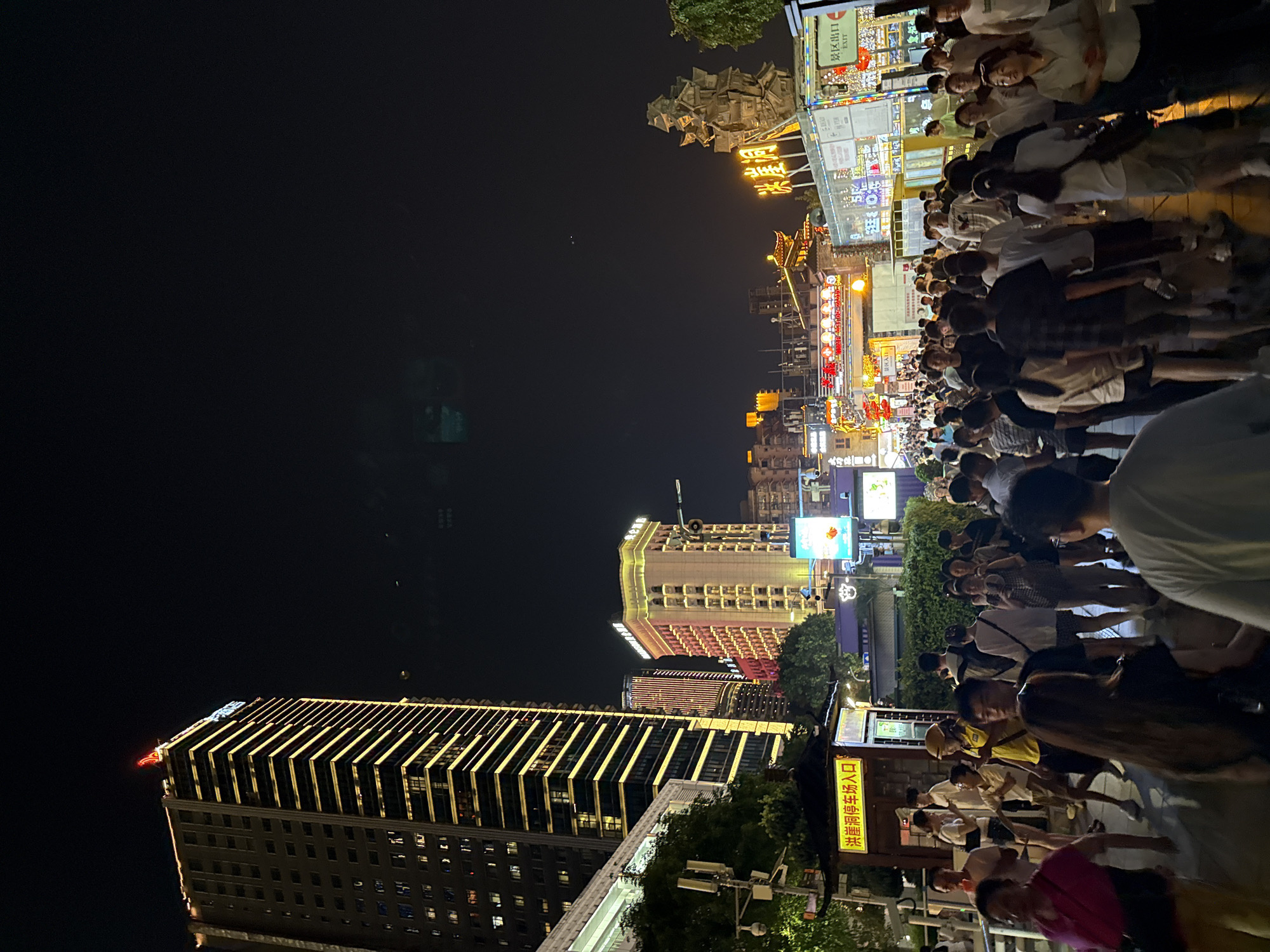 The image shows a crowded street scene at night in Yuzhong District, China. A tall, illuminated skyscraper dominates the left side of the frame, its facade lined with yellow lights. In the background, other buildings are visible, some with brightly lit signage and architectural details. The street is filled with a multitude of people walking in various directions, suggesting a lively commercial or entertainment area. Among the crowd, individuals are dressed in casual attire, and some are holding phones or bags. Signs in Chinese characters are displayed on buildings and kiosks, indicating the location is likely a commercial district or tourist area. The overall atmosphere is vibrant and bustling, illuminated by a variety of artificial lights against the dark night sky.