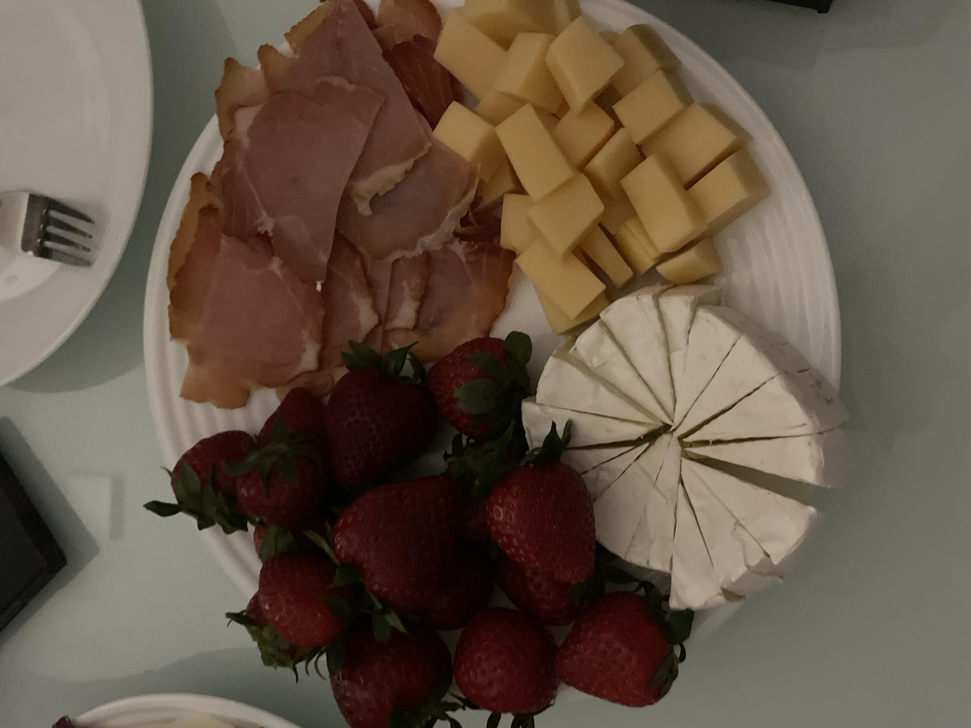 An overhead shot reveals a white oval platter laden with a variety of snacks, resting on a light blue-green surface. To the left side of the platter, a heap of thinly sliced, reddish-pink cured meat, resembling prosciutto, is neatly arranged. Adjacent to the meat, covering the upper right section, are numerous uniformly cut cubes of yellow cheese, likely a semi-hard type such as cheddar. In the lower right quadrant of the platter, a round, soft-rinded cheese, possibly brie or camembert, is cut into radial wedges. The bottom and lower-left areas of the platter are filled with fresh, bright red strawberries, many with their green caps still attached. A portion of a white ceramic plate with a silver fork is visible in the upper-left corner of the frame. The lighting is subdued, suggesting an indoor setting during the evening. There are no people or visible text. The platter's contents are arranged for a casual meal or snack in Novi Sad, Serbia.