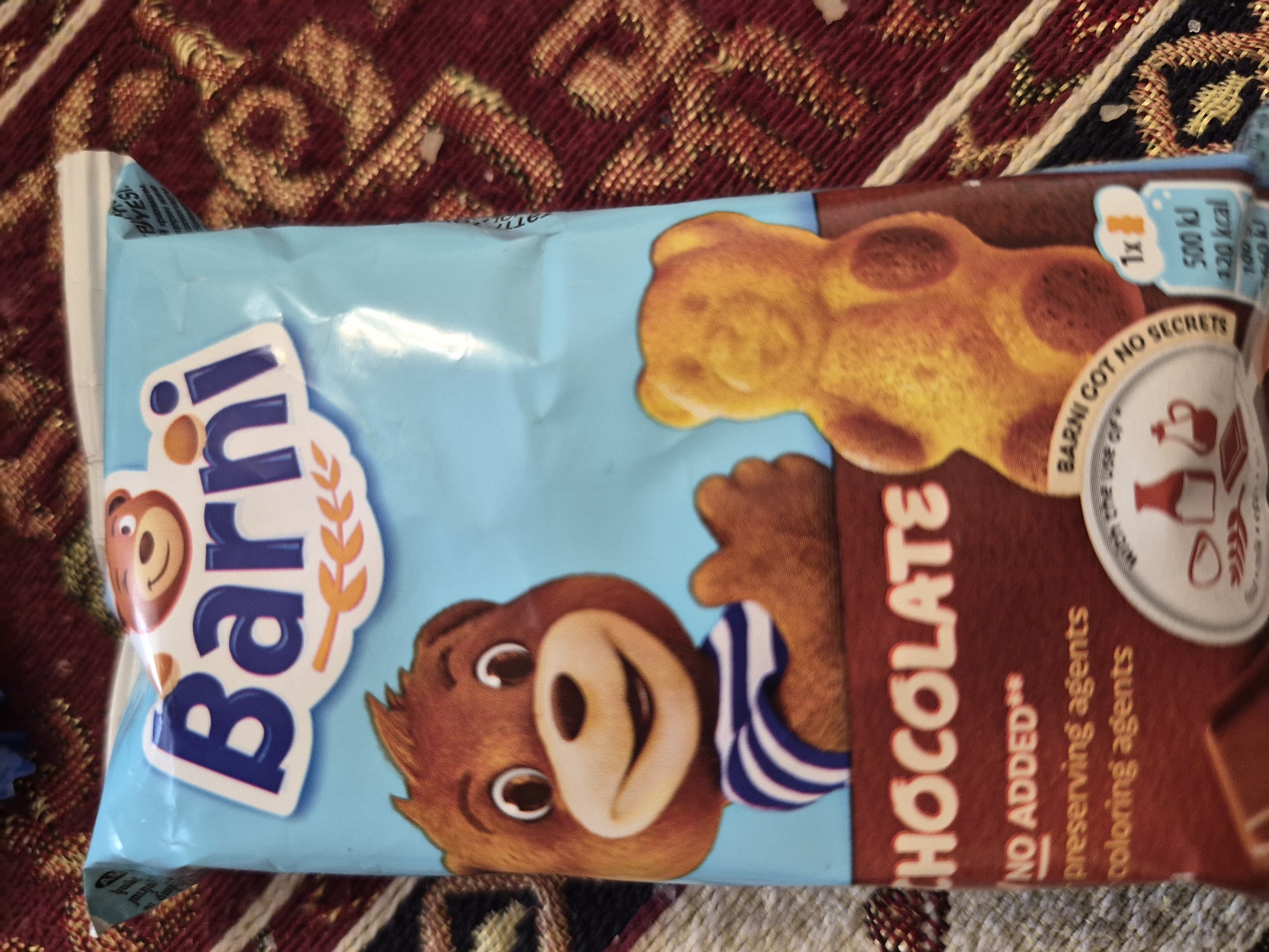 A package of Barni brand chocolate-flavored sponge cake snacks, featuring the brand's iconic bear character, is depicted resting on a patterned textile surface. The package, primarily light blue, is oriented diagonally across the frame, with its longer side extending from the top left to the bottom right. The design prominently features a friendly-looking cartoon bear wearing a blue and white striped shirt, along with a smaller golden bear-shaped cake, which is the product itself.
Visible text on the packaging includes the brand name "Barni" in white and dark blue letters, and the flavor "CHOCOLATE" in large red letters. Below the chocolate text, it states "NO ADDED** preserving agents coloring agents," indicating product claims. A circular emblem features the slogan "BARNI GOT NO SECRETS." Nutritional information for one serving (1x) is visible, showing "500 kJ" and "120 kcal." Faint text in the top left corner, possibly in a language other than English, includes "BIYESI," "И крахмаль," and "церин жан," which may relate to ingredients or other product information.
The textile surface beneath the package is a woven fabric with a warm color palette of deep red, gold, and cream, featuring intricate, possibly traditional, patterns. The overall scene suggests the item is casually placed, likely indoors in Yerevan, Armenia. There are no people visible, and no specific activity is taking place other than the presentation of the snack package.