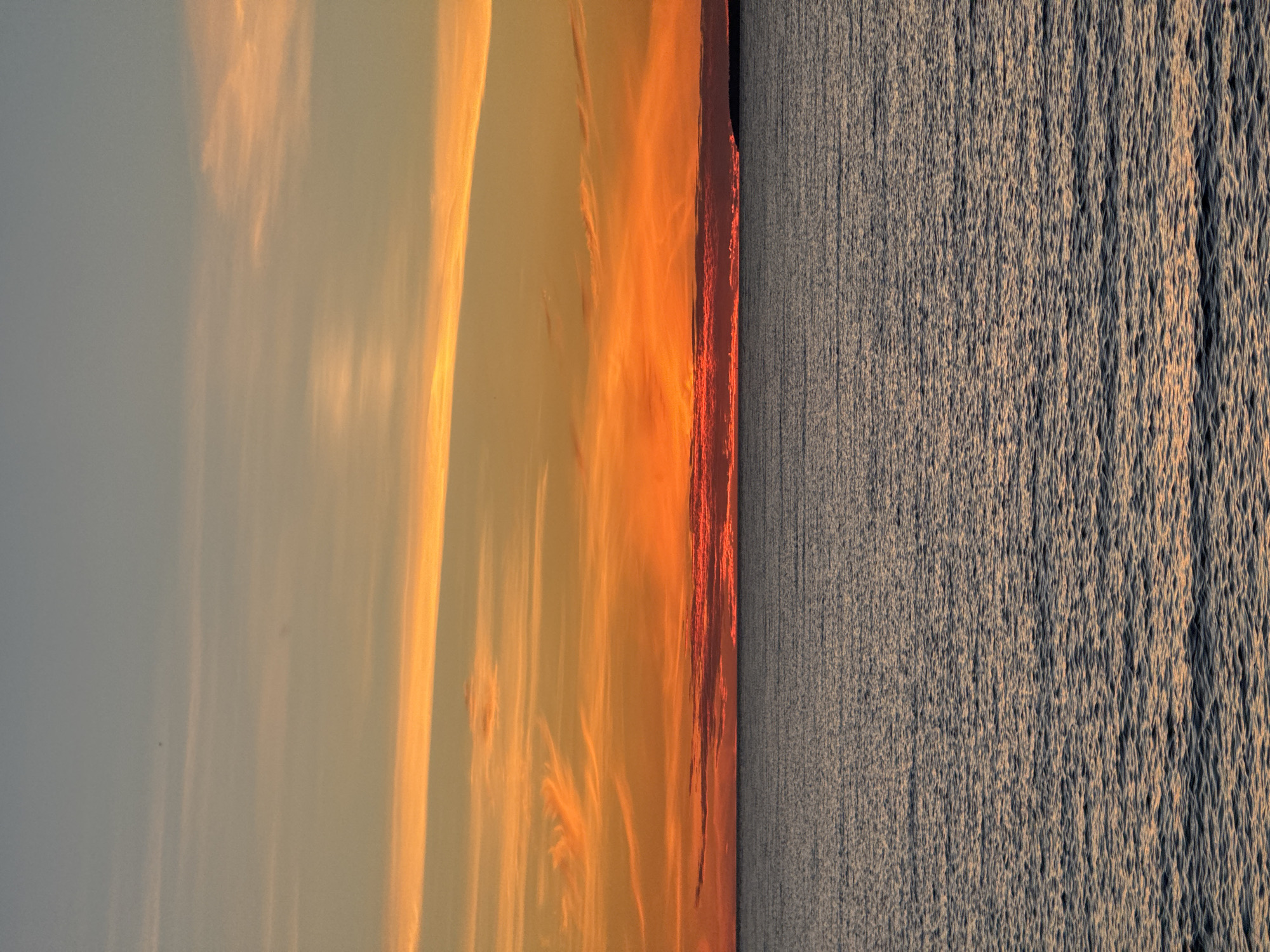 The image depicts a wide, horizontal view of a body of water under a vibrant sunset. The sky transitions from a pale blue at the top to warm oranges and reds near the horizon. wispy clouds are scattered across the sky, catching the sunset's light. A dark, silhouetted landmass is visible on the far right of the horizon. The water in the foreground is dark and textured with small ripples, reflecting the colors of the sky. There are no people, objects, or discernible actions in the scene. The location is not specified.