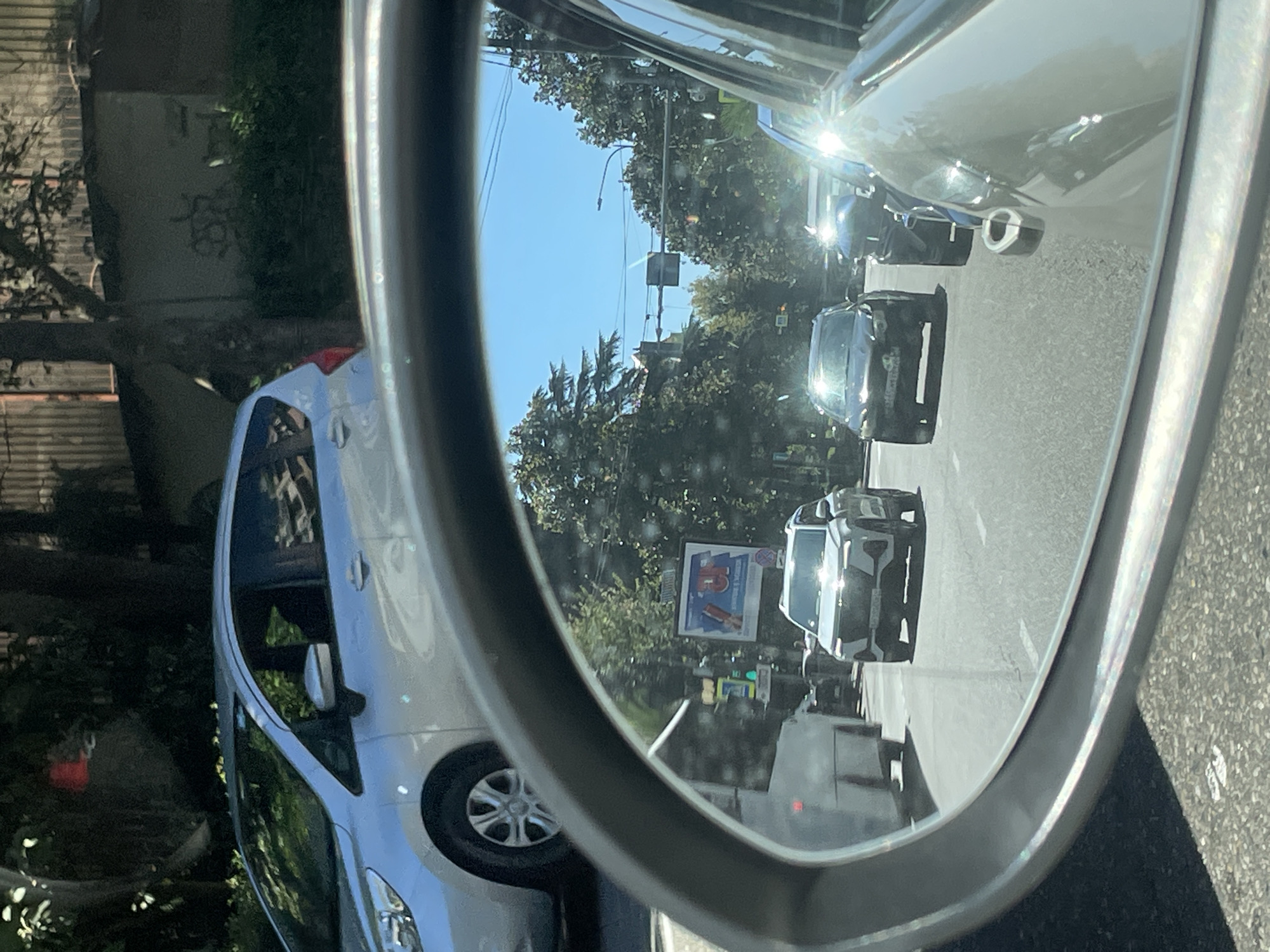 The image is a rear-view mirror reflection of traffic on a sunny day in Sochi, Russia. The reflection shows several cars, including a white SUV, a dark-colored car, and a silver minivan, all driving on a paved road lined with trees. A billboard advertising "39" is visible in the background. The sky is clear and blue. The original image shows a silver minivan from the side, parked on a street with trees and buildings.