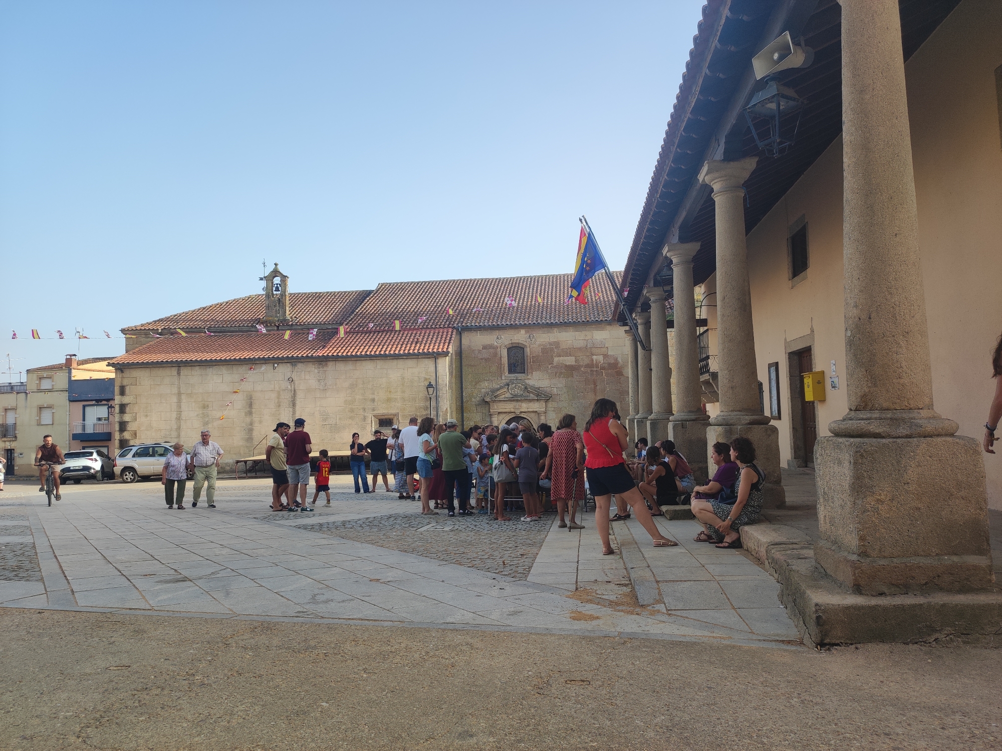In the city of Sobradillo, Spain, on a clear, sunny day, a plaza is depicted with a building featuring large, stone columns in the foreground, suggesting a public or historical structure. The building's facade is beige, with a tiled, terracotta roof. A flag, possibly a regional or organizational one with blue, yellow, and red elements, is visible attached to the structure. In the mid-ground, a church or similar religious building with a red-tiled roof and a bell tower is present. Numerous people are gathered in the plaza, some standing in groups, others walking or sitting. Several individuals are conversing, while others appear to be observing or waiting. A man is riding a bicycle on the paved plaza. Cars are parked in the background, indicating it's an urban setting. Small flags or banners are strung across the plaza and on the church building. The ground surface is a mix of large stone tiles and compacted dirt.