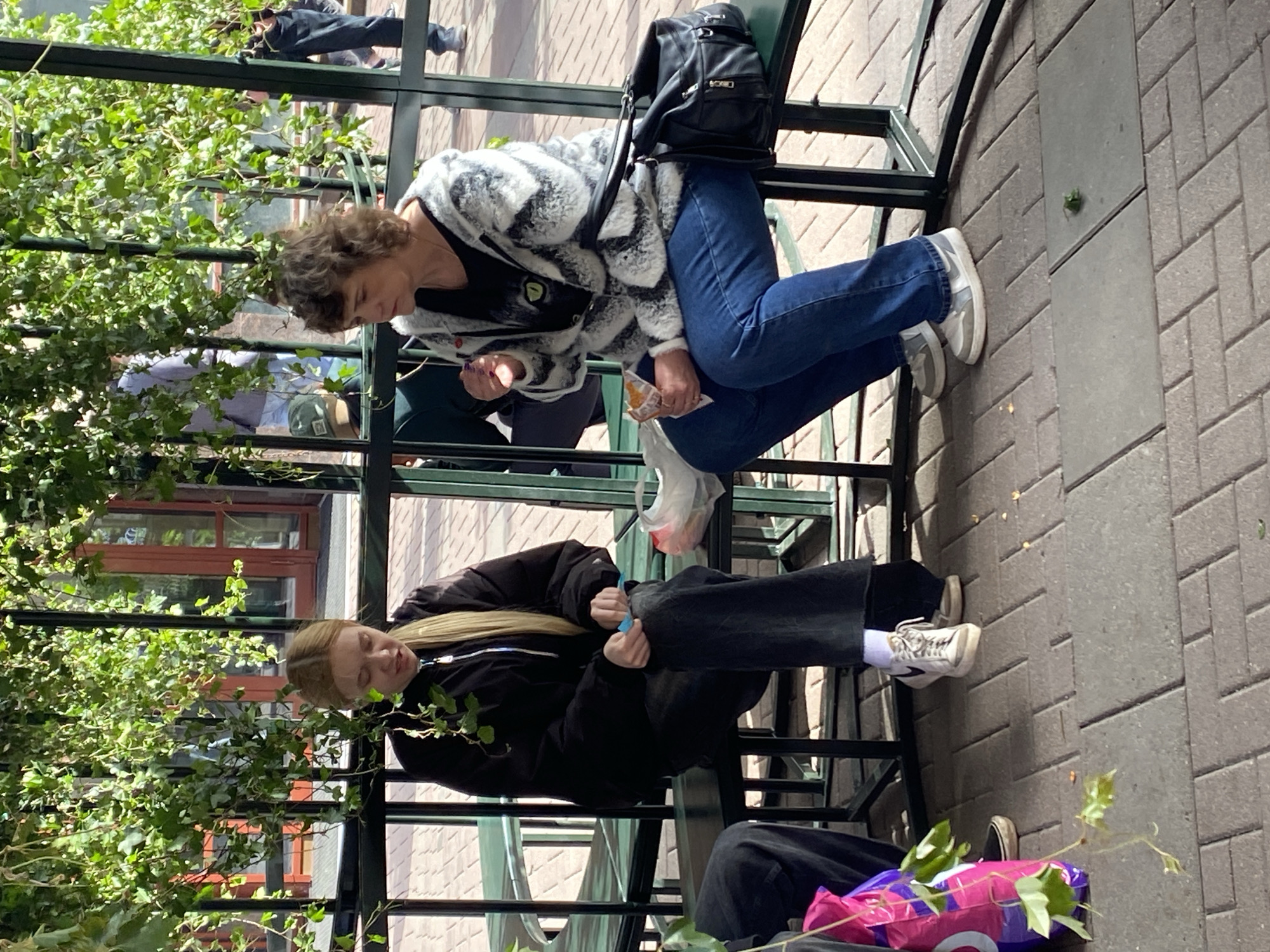 Two women are sitting on adjacent benches in an outdoor area. The woman on the right is wearing a black and white patterned jacket, blue jeans, and gray sneakers. She has a black backpack on the back of her chair and is holding a package in her hand. The woman on the left is wearing a black jacket, black pants, and white sneakers. She appears to be opening a bag of some kind. The benches are black metal and are partially covered with greenery. The ground is paved with light gray bricks. The background shows a building with red-framed windows.
It is not possible to confirm this image is in Moscow, Russia.