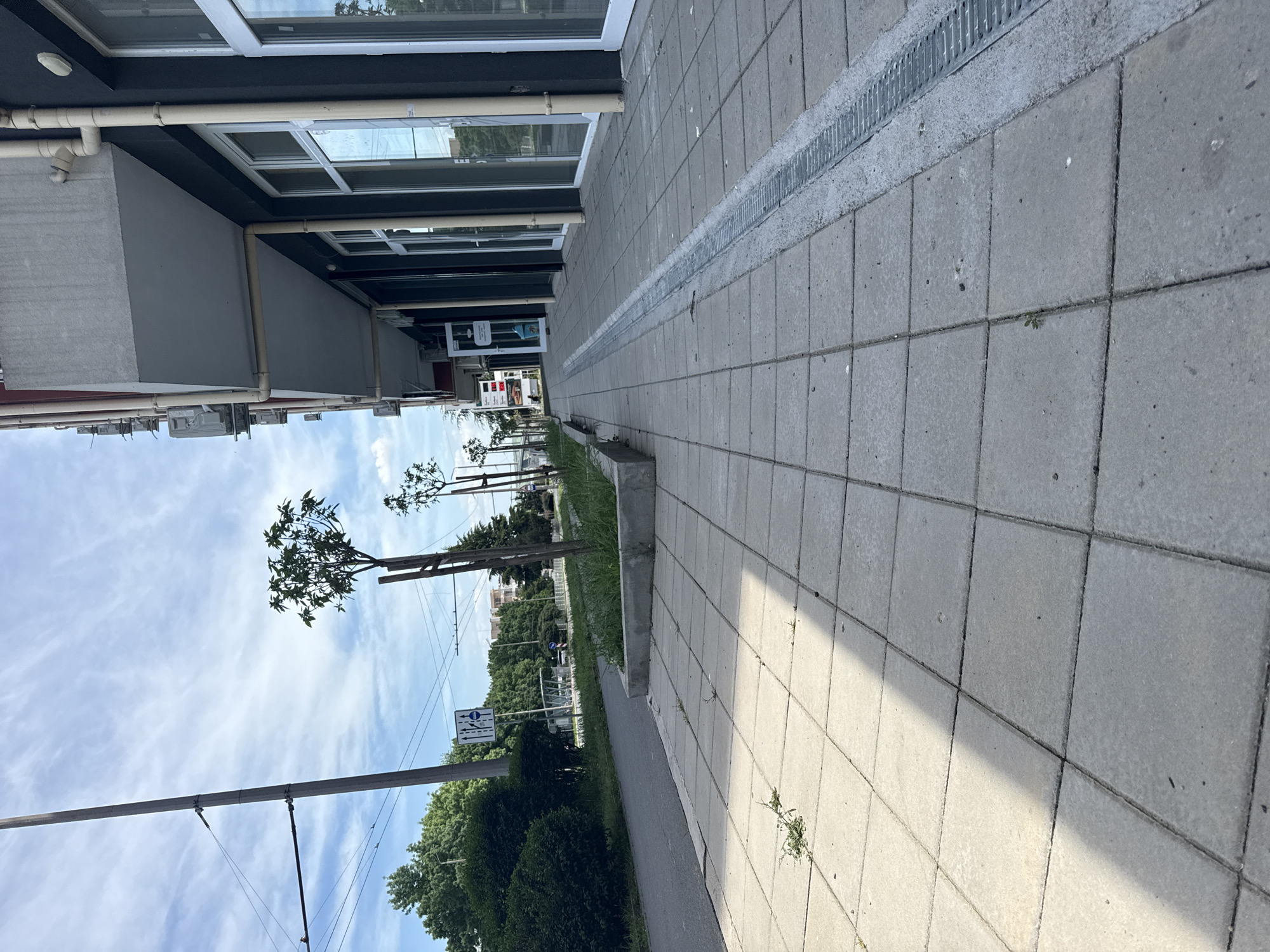 The image shows a sidewalk in Burgas, Bulgaria. The sidewalk is made of square concrete tiles and is adjacent to a building on one side and a green area with trees on the other. A metal grate runs along the edge of the sidewalk next to the building. Overhead electrical wires are visible against a sky with some clouds. The perspective is slightly tilted.