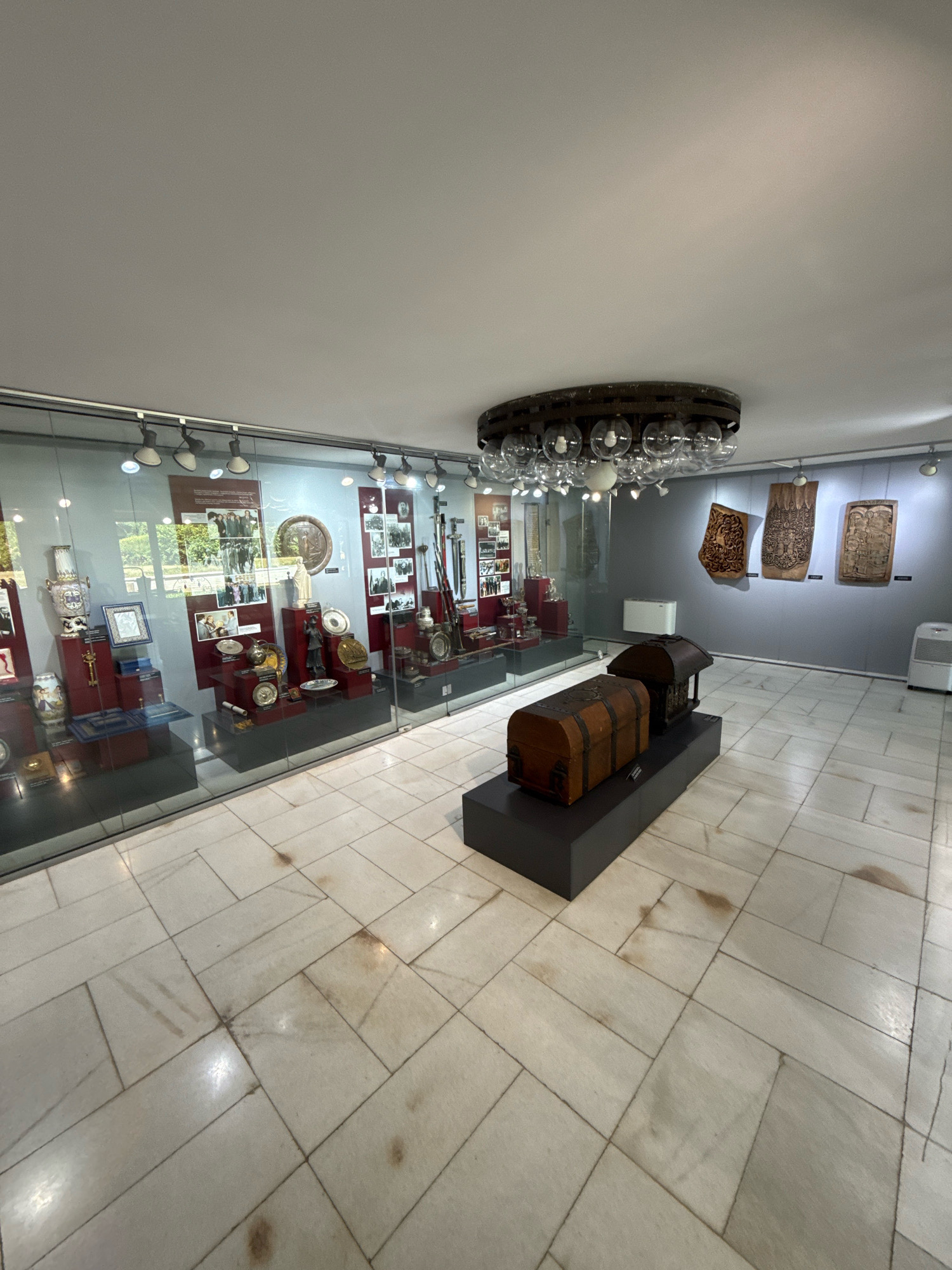 This media file depicts an indoor exhibition space, likely a museum or gallery, with a polished marble floor. The setting appears to be a well-lit indoor environment, possibly during the day, given the ambient lighting. The primary subjects are various historical artifacts displayed behind glass or on pedestals.
In the foreground, two wooden chests are prominently displayed on a dark, raised platform. One chest is larger and positioned in front of a smaller, more ornate one. To the left, a long glass display case showcases a collection of smaller items, including vases, framed pictures, plates, and decorative objects, arranged on red and blue pedestals. The reflection in the glass reveals an outdoor scene with greenery, suggesting the museum might be situated in a park or has windows looking out onto one.
Further into the space, a large, circular chandelier with multiple glass bulb-like fixtures hangs from the ceiling. The wall in the background is painted a neutral gray and features several large, carved wooden panels with intricate designs, hung at eye level. White air conditioning units are visible on the wall and floor.
The overall atmosphere is one of quiet contemplation and historical preservation. There is no visible text in the image. The location context of Pravets, Bulgaria, suggests these artifacts might be related to the history or culture of that region. No people are visible in the media file.