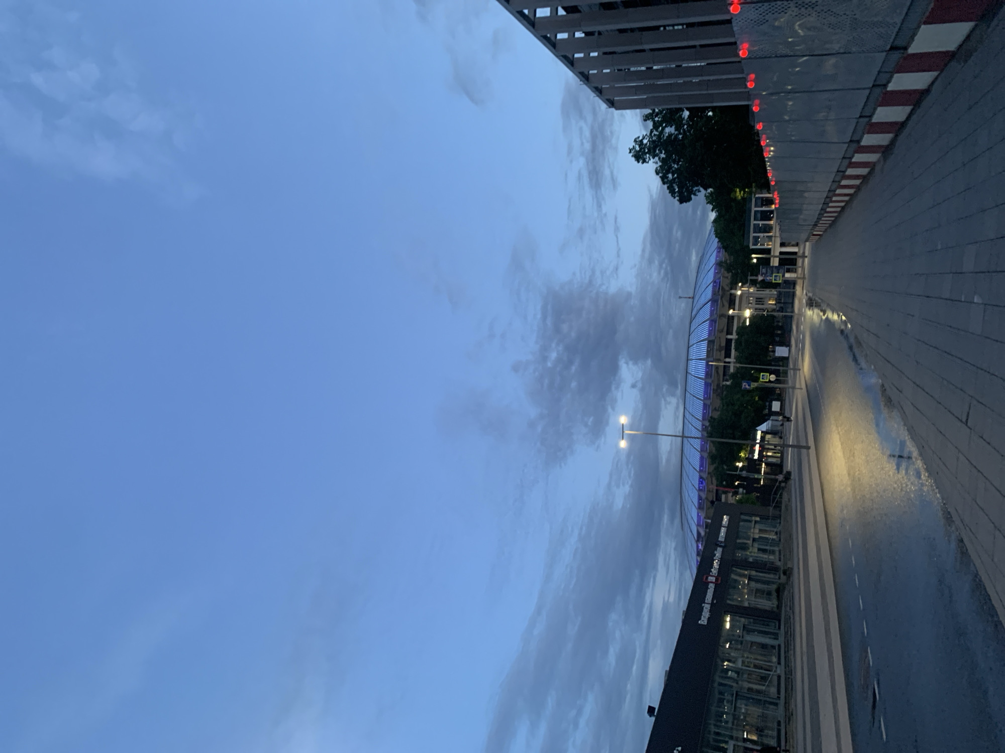 The photograph captures an urban scene under a dimly lit sky. The sky is dominated by shades of blue, ranging from a lighter hue to a deeper twilight tone, with scattered clouds adding depth to the atmosphere.
On the right side of the frame, a paved walkway stretches into the distance. The walkway appears wet, with light reflecting off the damp surface, suggesting recent rainfall. Adjacent to the walkway is a barrier or edge, marked with a pattern of alternating red and white stripes, possibly indicating a restricted area or boundary.
To the left, a modern building with a dark facade and reflective windows stands tall. The building's architecture is angular, with a mix of solid walls and glass panes. A few visible signs in Cyrillic script adorn the building, hinting at its location within a Russian-speaking region. The presence of streetlights along the walkway and within the building premises indicates the scene is set at dusk or night. A sports stadium is visible in the background.