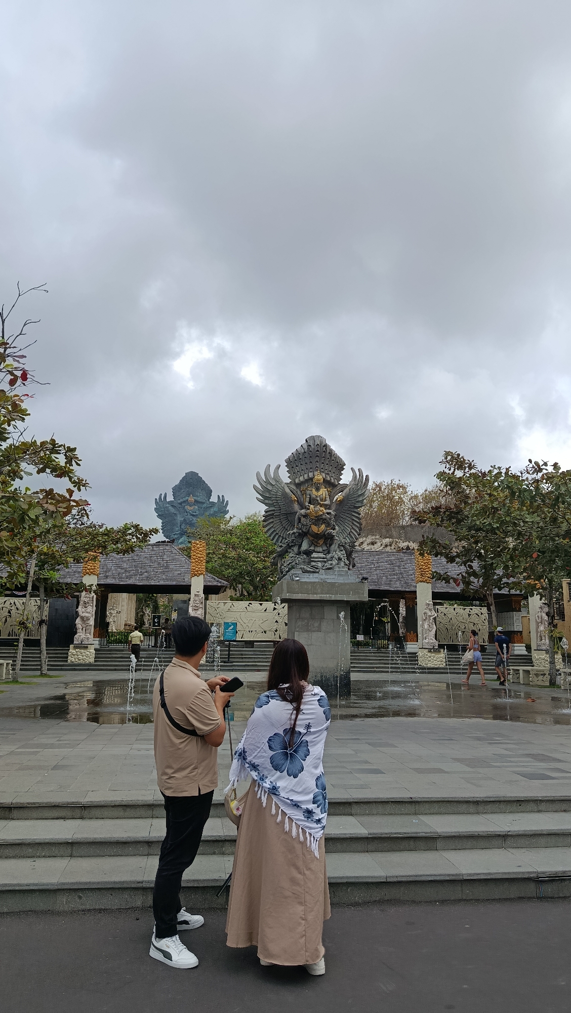 This is a vertical, eye-level shot of the Garuda Wisnu Kencana Cultural Park in Kuta Selatan, Indonesia, on an overcast day. In the foreground, a man and a woman stand with their backs to the camera, facing a large bronze statue of Garuda, a mythical bird-like creature. The man, wearing a beige polo shirt and black pants, holds a phone in his left hand. The woman, in a long beige dress and a white shawl with blue floral patterns, stands beside him. To the left of the Garuda statue, another large blue statue, likely also Garuda, is visible in the distance. In front of the statues, a fountain sprays water. The park features traditional Balinese architecture with wooden roofs and stone carvings. Several other people are seen walking in the background, near the entrance to the park.