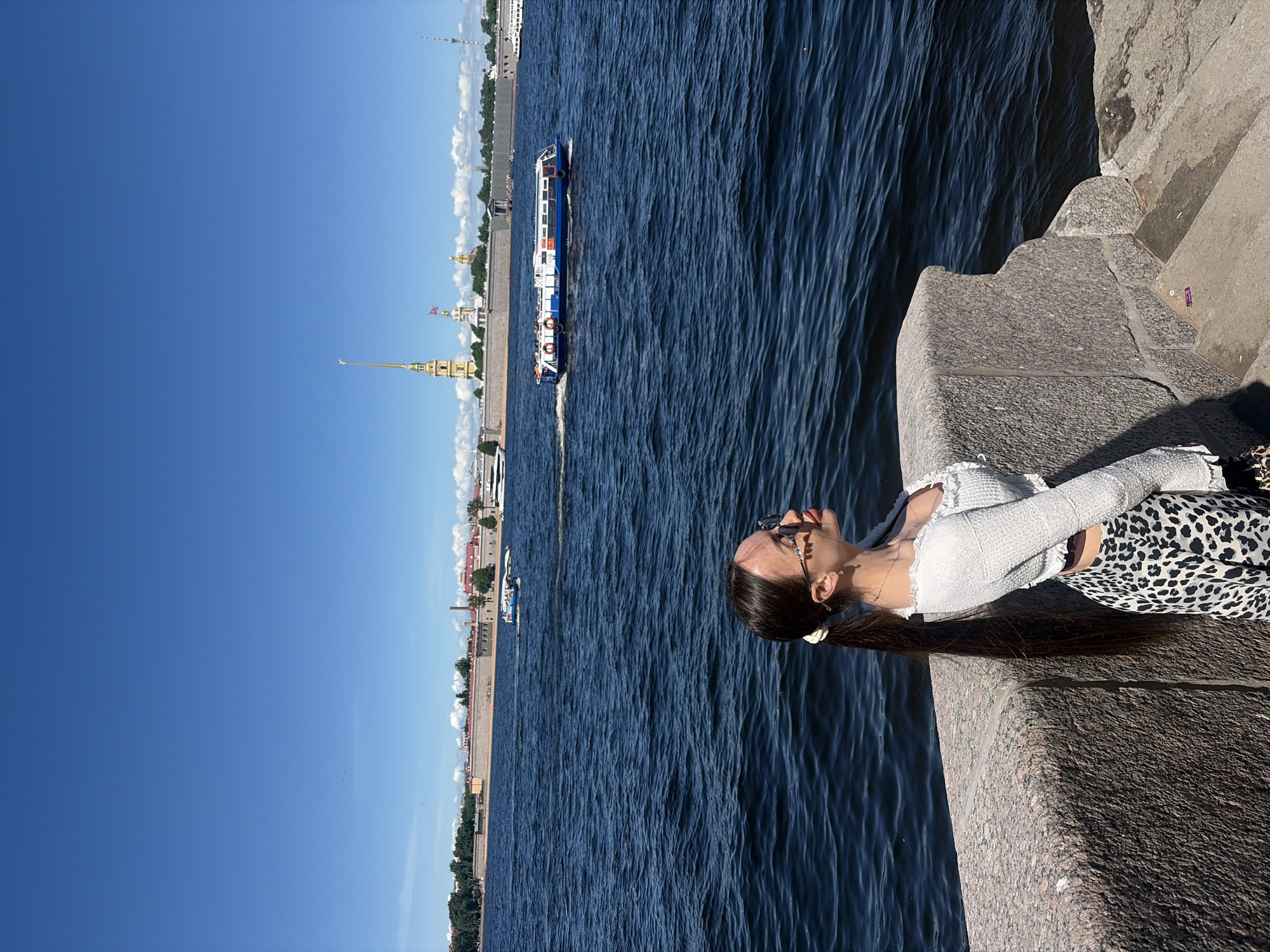 The image portrays a sunny daytime scene along the Neva River in Saint Petersburg, Russia. The background features the Peter and Paul Fortress with its iconic golden-spired Peter and Paul Cathedral and other associated structures. The sky is clear blue with scattered white clouds near the horizon.
In the foreground, a young adult female is positioned on the right side, leaning against a sturdy, grey granite embankment wall. She has long, dark brown hair tied in a low ponytail with a white scrunchie, extending well past her waist. She wears dark sunglasses, a white, long-sleeved, textured crop top, and a black and white leopard-print midi skirt. Her head is tilted upwards and to her right, away from the camera, with a subtle expression showing her tongue slightly protruding.
The Neva River, dark blue and subtly choppy, occupies the midground. A blue and white passenger boat, with orange lifebuoys visible, moves from left to right across the water, leaving a visible white wake behind it. Other smaller vessels are discernible in the distance. To the far right, a section of granite steps descends from the embankment towards the water, with a small, discarded purple wrapper lying on one of the steps.