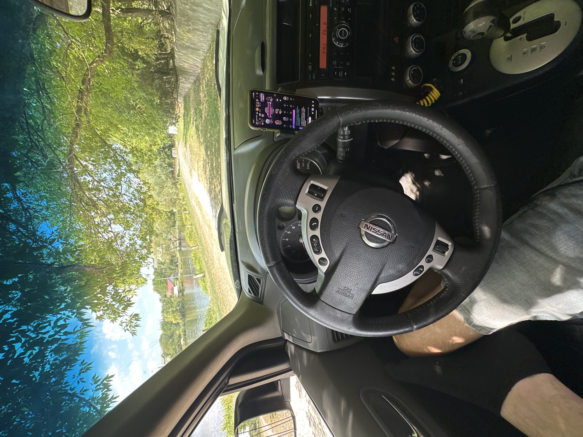 The image shows the interior of a Nissan SUV parked by a lake on a sunny day. The view is from the driver's seat, looking out through the windshield and side window. The steering wheel, dashboard, and center console are visible. A smartphone is mounted on the dashboard, displaying a social media app. Through the windshield, a calm body of water, trees, and houses can be seen. The location is identified as Makiv, Ukraine. The scene appears peaceful and scenic, with the car parked in a spot overlooking the lake.