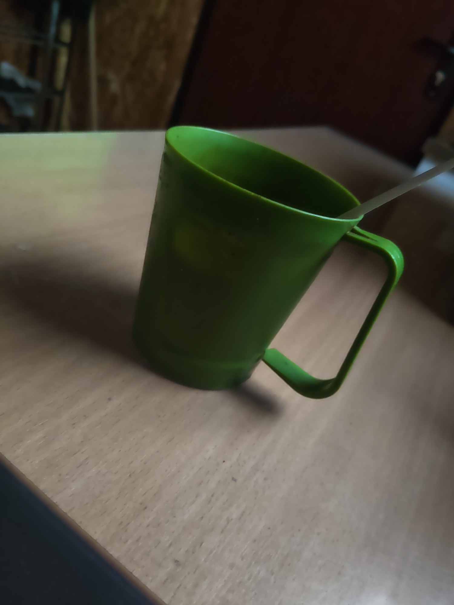 A green plastic mug with a white spoon rests on a light brown wooden table. The mug is positioned at an angle, with its handle facing right. The background is blurred, showing what appears to be a wall with textured panels. The scene is lit from the left, casting a shadow to the right of the mug. There are no people visible in the image. The location is identified as Platoshino, Russia.