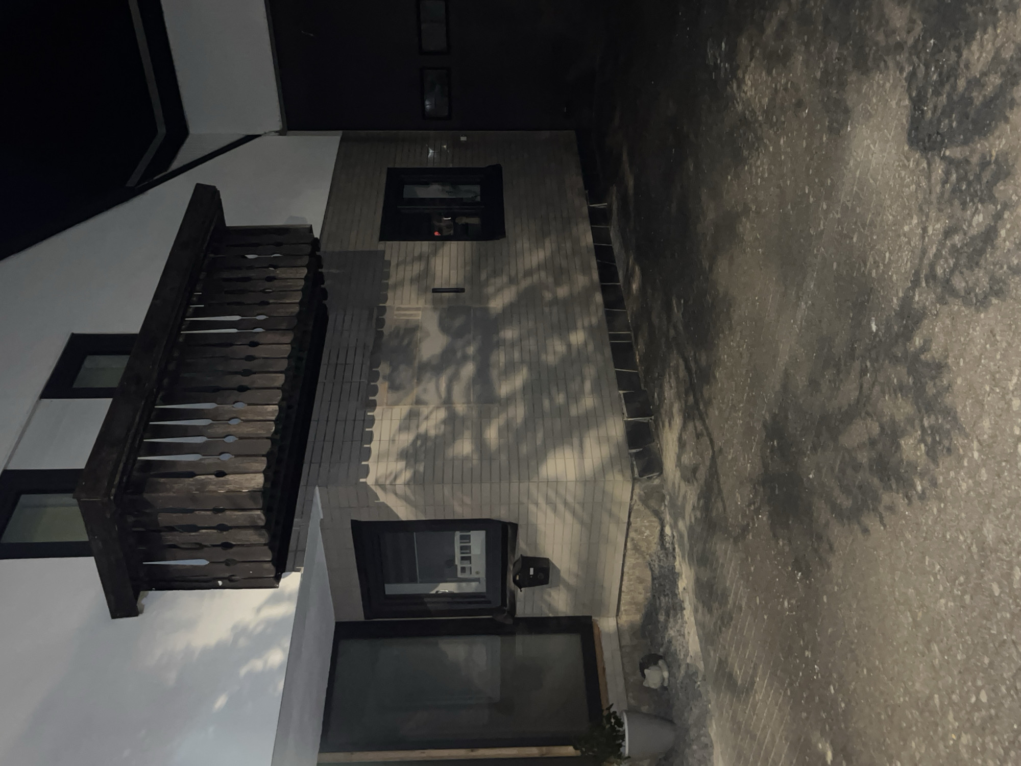 This is a night photograph of a modern house in Saint-Vith, Belgium. The house has a white facade and a dark-colored, wooden balcony with ornate railings. A section of the house is constructed with light-colored brick. There are several windows visible, some with dark frames. One window reflects a white building. A black mailbox is mounted on the brick wall below a window. A potted plant is situated on the ground near a large glass door or window on the left side of the image. The ground in front of the house appears to be paved with rough, grey material, possibly asphalt or concrete, with some gravel and debris scattered on it. Shadows of tree branches are cast across the brick wall and the ground, illuminated by an unseen light source. To the right of the house, a dark, unlit area, possibly a garage or an extension, is visible, with rectangular windows on its facade. The overall impression is one of a residential property at night.
