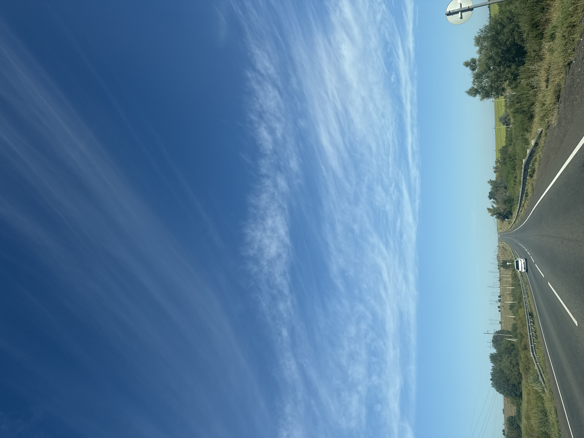The image depicts a clear, sunny day on a straight, empty highway in Gamaleevka-1, Russia. The road stretches into the distance, bordered by green fields and scattered trees on either side. A white car is visible in the distance, driving in the same direction as the camera. The sky is a vibrant blue, with wispy white clouds streaking across it. The overall impression is one of open space and a peaceful, rural landscape.