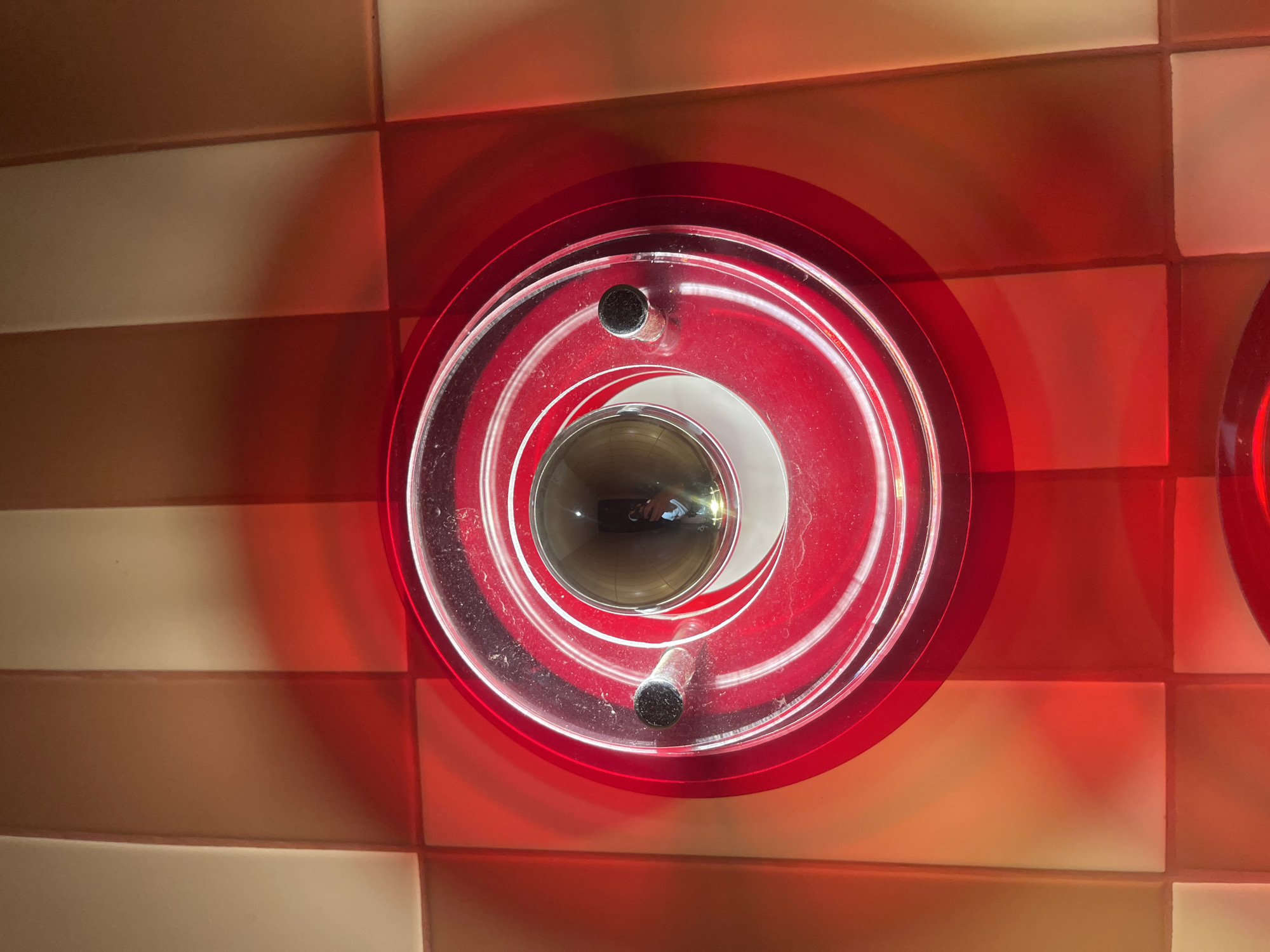 A low-angle, eye-level photograph reveals a circular, multi-layered, translucent red plastic object mounted on a tiled wall. The tiles are arranged in alternating rows of light beige and brown. The object consists of several concentric rings that converge towards the center, creating a tunnel effect. Two black cylindrical objects attach to the inner rings of the plastic fixture. Reflections inside the central opening suggest the photographer and some of the surrounding room. The edge of another, similar object is visible on the right side of the frame. The location given is Bac Nha Trang Ward, Vietnam.