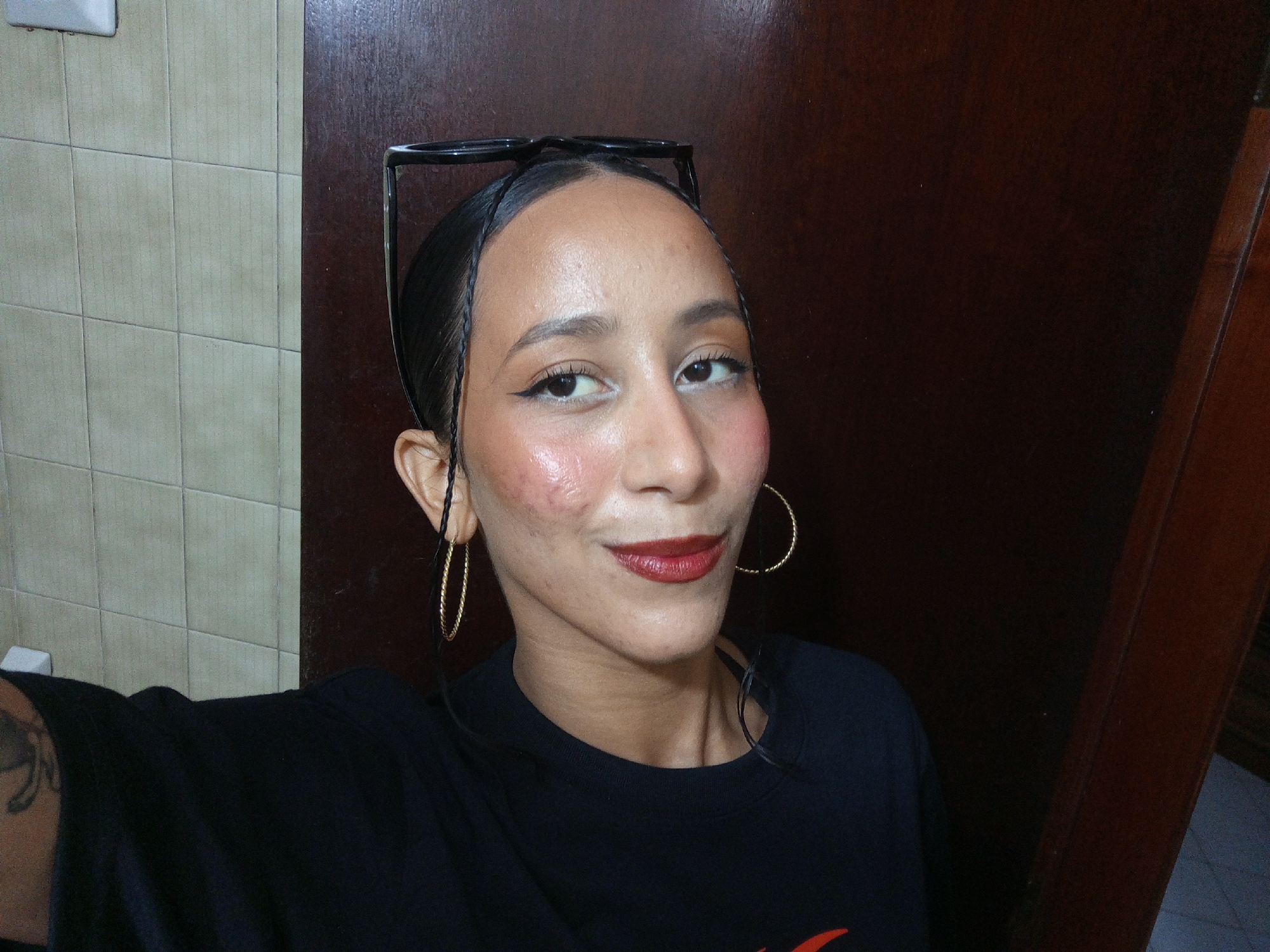 A person with light brown skin, red lipstick, and gold hoop earrings poses for a selfie. The person wears a black t-shirt. A pair of black glasses are perched on top of the person’s head. The person is positioned in front of a wall with light-colored tiles and a dark brown door or wall. A tattoo is visible on the person’s left arm. It is not possible to determine the exact location or city from the image.