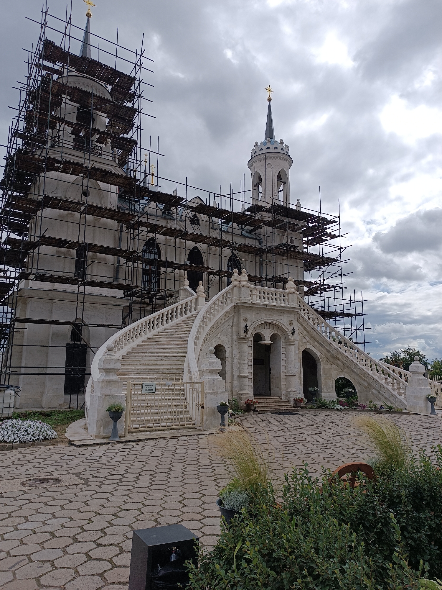 The image shows the Church of the Vladimir Icon of the Mother of God in the village of Bykovo, Russia, undergoing restoration. The church's facade and towers are extensively covered with scaffolding, indicating ongoing construction work. A grand, ornate white stone staircase leads to the main entrance. The sky is overcast with thick clouds. The foreground features a cobblestone courtyard with scattered decorative elements like potted plants and greenery. The architectural style appears to be neo-Gothic or classical with some baroque influences. No people are visible in the frame. The overall scene suggests a historical landmark being preserved.