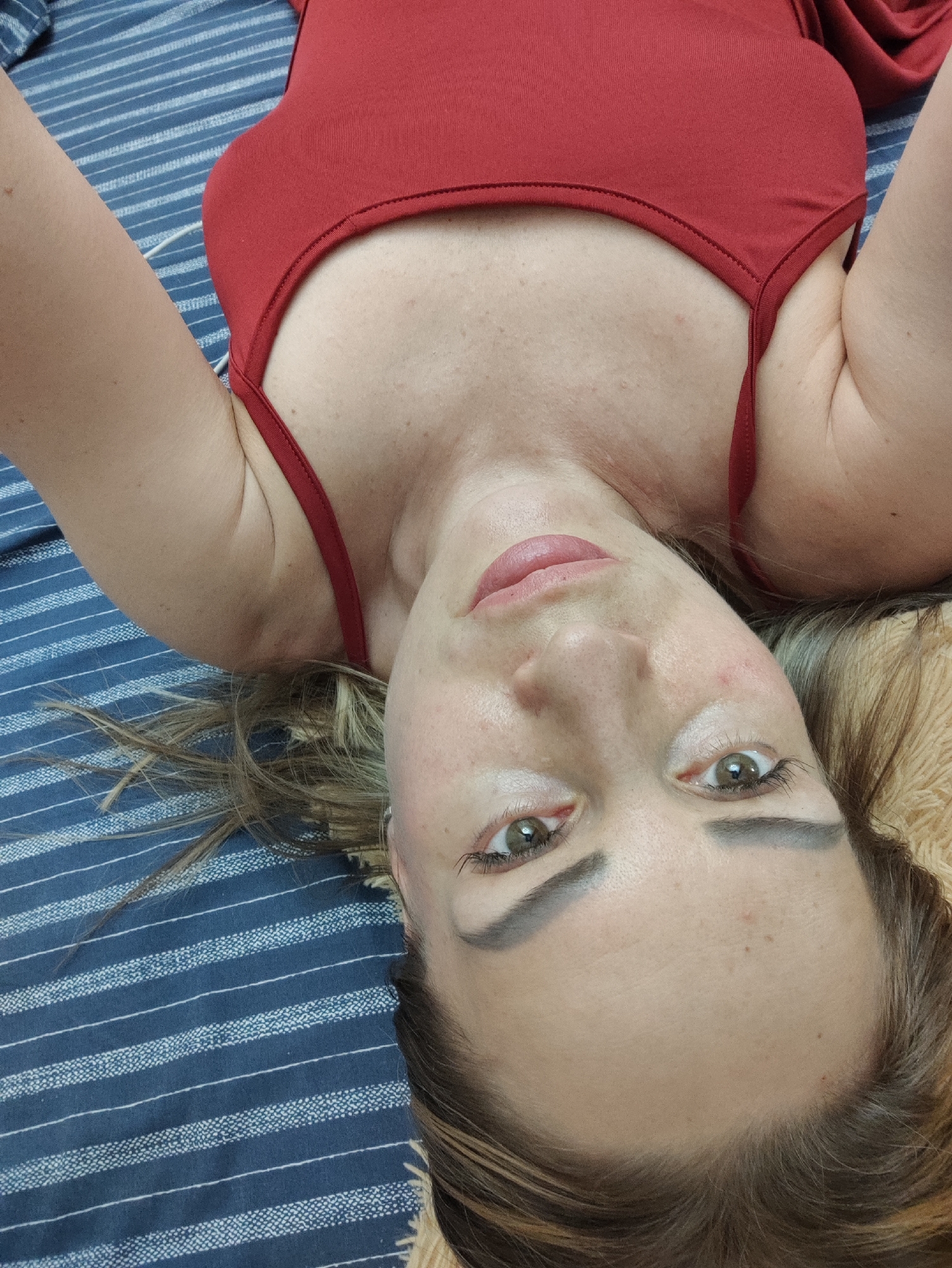 This is a close-up, upside-down selfie of a woman lying on a blue and white striped surface, likely a bed or couch. She is wearing a red sleeveless top. Her face is angled towards the camera, with her eyes open and looking directly at it. Her hair, a mix of blonde and brown, is spread out around her head. Her makeup includes mascara and eyeliner. The lighting appears to be indoor. The scene is a personal, casual setting. There is no visible text. The location is specified as Almaty, Kazakhstan, but this context is not visually evident in the image itself.