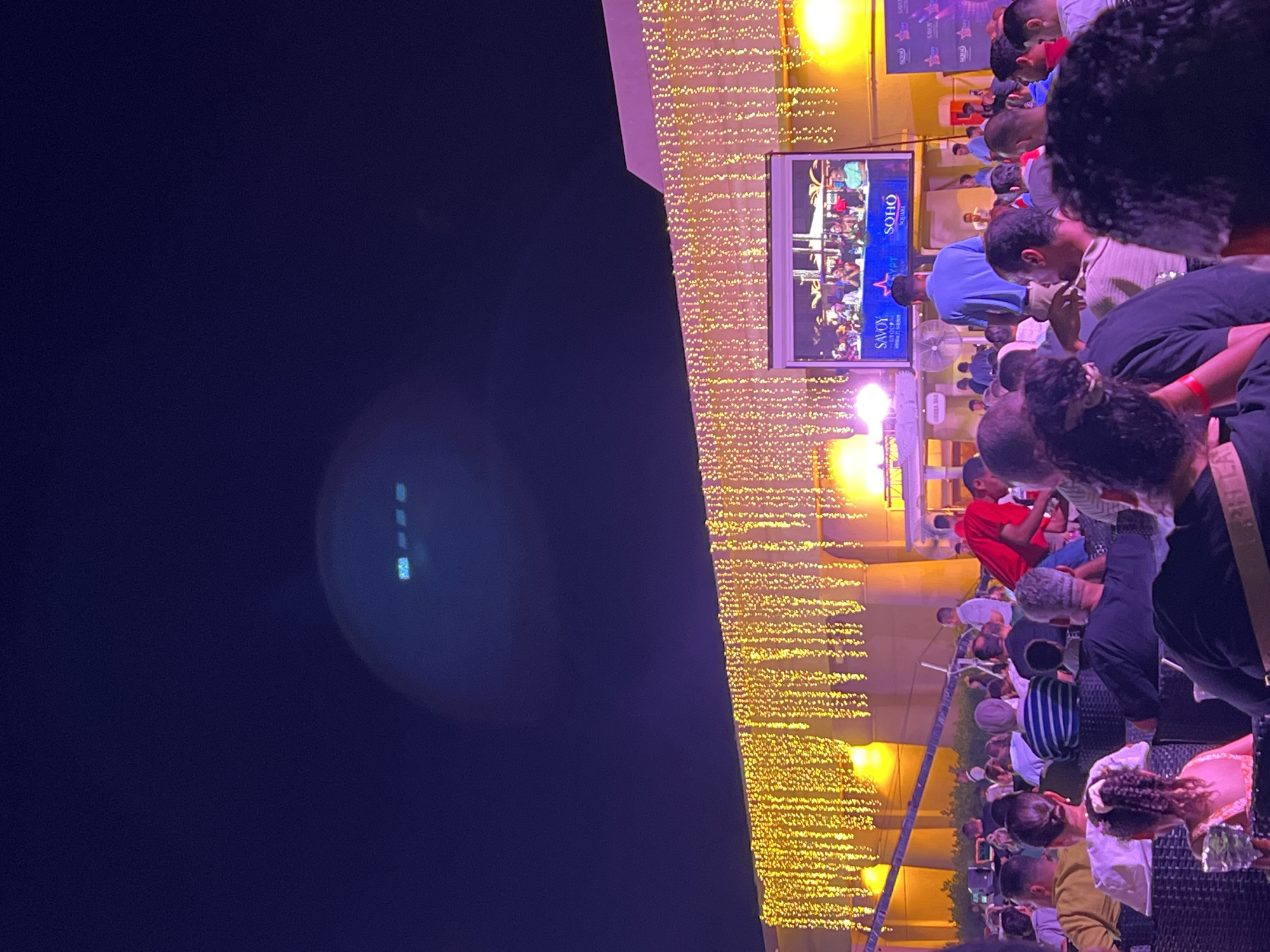 This image depicts an outdoor evening event at Soho Square in Sharm El Sheikh, Egypt. A large crowd of people is gathered, seated in an outdoor seating area, facing a large screen. The screen displays a panel of individuals sitting behind a table, with logos for "SAVOY" and "SOHO SQUARE" visible. The building behind the screen is decorated with numerous hanging strings of golden lights. The sky above is dark, indicating nighttime, and there is a bright lens flare or light artifact in the upper portion of the sky. The overall atmosphere suggests a public viewing or performance event.