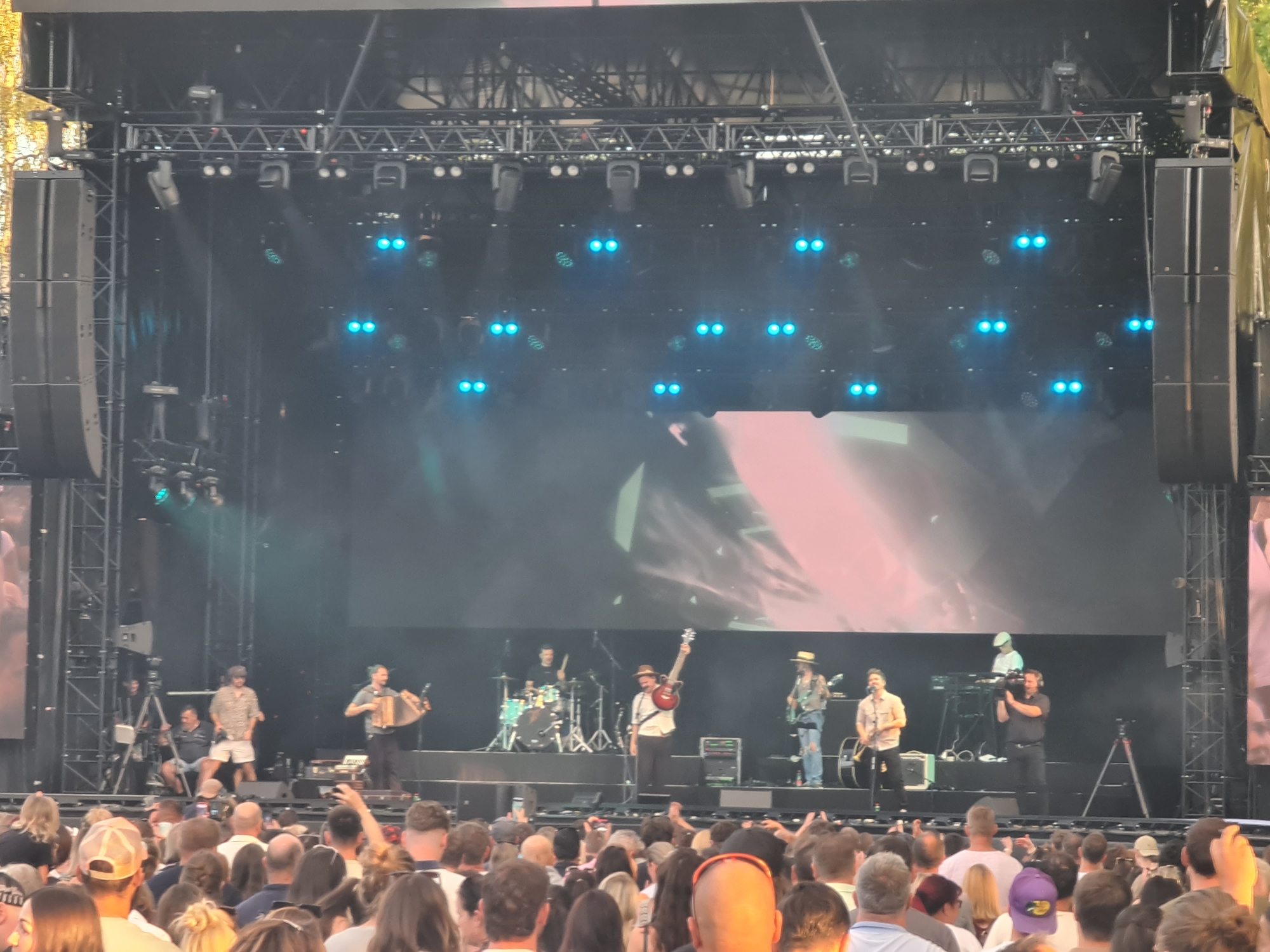 This is a photograph of a live band performing on an outdoor stage in Klagenfurt, Austria. The stage is set up with professional lighting and a large screen in the background displaying abstract imagery. A band of at least six musicians is on stage, playing various instruments including an accordion, drums, and guitars. Some of the musicians are singing into microphones. There are several crew members on stage, including a person operating a video camera and others managing equipment. In front of the stage, a large crowd of spectators is gathered, many of them holding up their phones to record the performance. The overall atmosphere appears to be that of a lively outdoor concert. The time of day appears to be late afternoon or early evening, as the lighting on stage is primarily artificial.