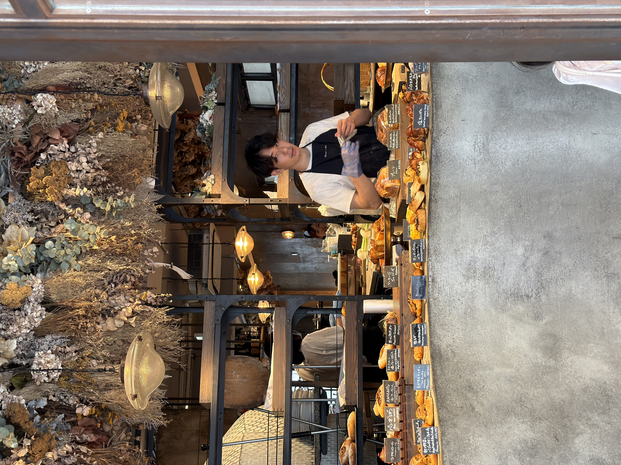 The image depicts the interior of a bakery in Meguro, Japan, with a focus on the bread display and a young male staff member.
The bakery has a rustic and natural aesthetic. Dried flowers and foliage are densely hung from the ceiling, creating an organic canopy. Two vintage-style pendant lights with glass shades hang from the ceiling, emitting a warm glow.
In the foreground, a display counter showcases a variety of baked goods, including loaves of bread, pastries, and rolls. Many of these items have small black signs with Japanese text and prices. The counter itself is made of a textured, grey material resembling concrete.
A young man, likely a baker or staff member, stands behind the counter. He is wearing a white t-shirt and a black apron, and has blue disposable gloves on his hands. He is holding a white cloth and appears to be wiping his hands or the cloth. His expression is neutral as he looks towards the camera.
Behind the counter, a multi-tiered shelving unit holds more bread and some other items, including what looks like plates or bowls on the left shelf. In the background, other staff members can be seen, including a woman in a light-colored top visible on the left, and another person with dark hair partially visible on the far right.
The overall impression is that of a welcoming and artisanal bakery. The abundance of dried plants and the display of fresh bread contribute to a sensory experience, suggesting a focus on quality and natural ingredients. The setting in Meguro, a district known for its trendy boutiques and cafes, aligns with this impression.