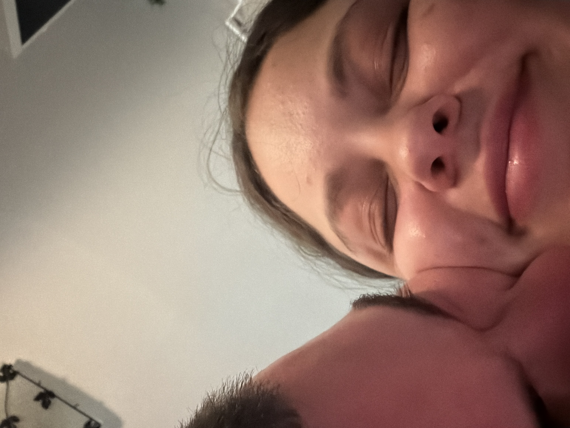 The image shows two people in close proximity. One appears to be a woman with her eyes closed and a slight smile, positioned closer to the camera. The other person, likely a man, is partially visible with dark hair and their face nuzzled against the woman's face. The background is a blurry, light-colored wall. The location in Varna, Bulgaria is not discernible from the image.