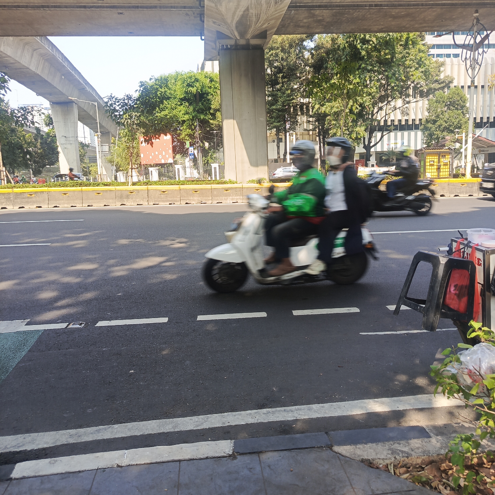 The image captures a street scene in South Jakarta, Indonesia, under an elevated concrete structure, likely an overpass or bridge. In the foreground, a white scooter is moving rapidly from left to right, with its two occupants blurred due to motion. The rider wears a green jacket and a helmet, while the passenger behind them is dressed in dark clothing and also wears a helmet. The scooter is on an asphalt road marked with white lines. To the right of the scooter, a black plastic chair and a small cart with red and white containers are visible on the side of the road. In the background, trees and buildings are present, along with other vehicles in motion, indicating traffic. The scene is brightly lit by sunlight, casting shadows on the road.