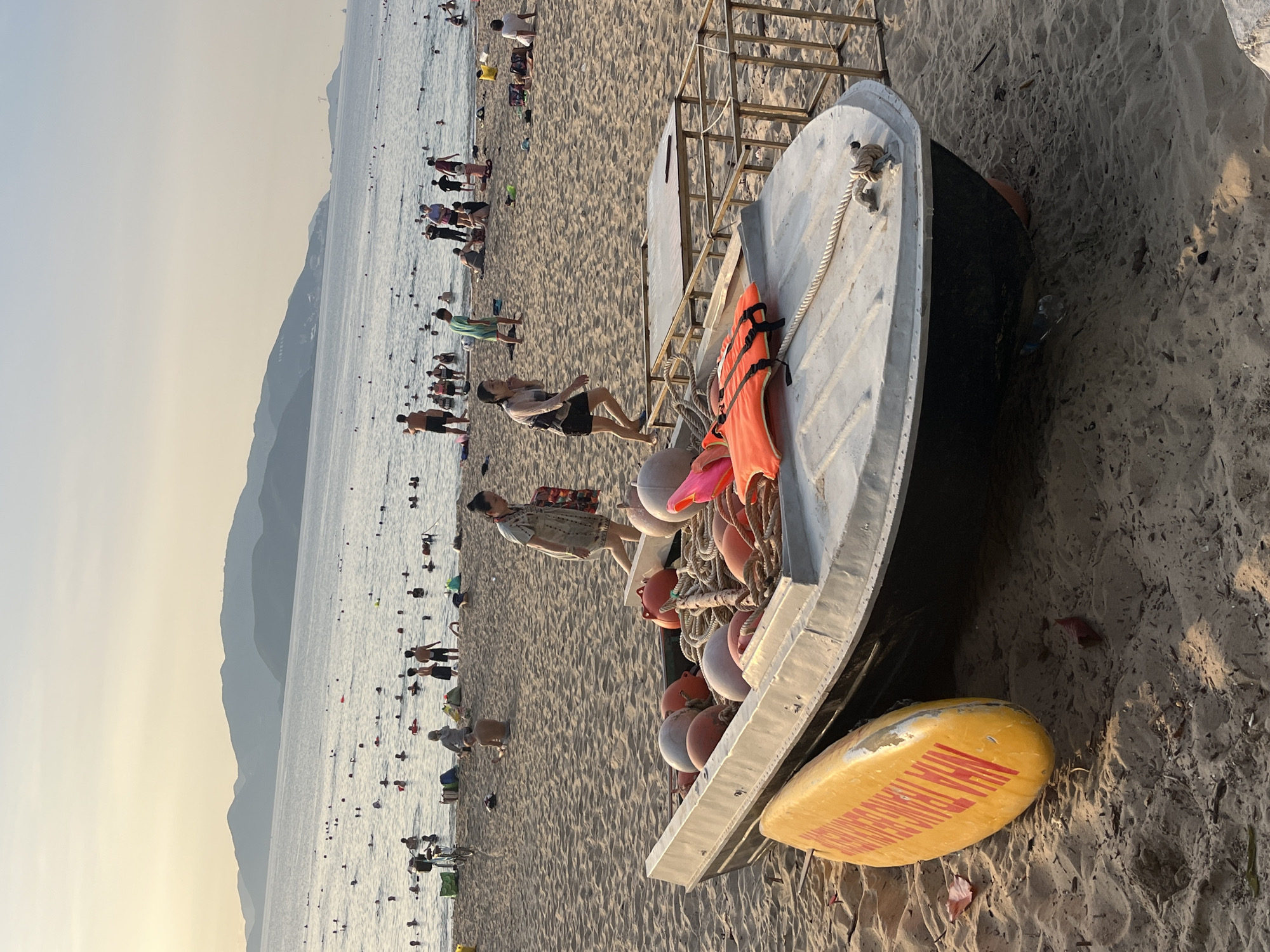 The image shows a crowded beach in Nha Trang Ward, Vietnam. The beach is sandy and slopes down to the sea, where many people are swimming and playing. In the foreground, a small boat sits on the sand, laden with orange life jackets and buoys. A yellow surfboard rests beside the boat. People are walking and standing on the beach near the boat. Hills are visible in the background. The light suggests it is late afternoon or early evening.