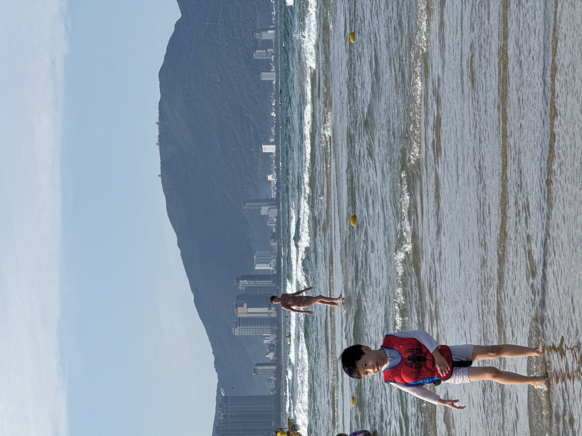 This image depicts a sunny beach scene in Da Nang, Vietnam, showcasing individuals enjoying the ocean against a backdrop of urban development and a large mountain. The photo is oriented vertically, but when viewed horizontally, the subjects stand upright.
In the immediate foreground, a young boy is standing in shallow water, looking towards the viewer with a slight smile. He wears a red and blue life vest featuring a black Spider-Man logo, a light-colored long-sleeved shirt, and light shorts. His feet are partially submerged in the clear water. Further back, another person, likely a woman, is standing in the water up to her waist, wearing a light-colored two-piece swimsuit and facing away from the camera, towards the open sea.
The ocean features gentle waves breaking towards the sandy shore. Two bright yellow buoys are visible in the water, indicating a designated area. Along the coastline, numerous multi-story buildings, likely hotels or residential blocks, stretch along the base of a vast, dark green mountain that dominates the left side of the frame. The sky above is clear and bright blue with minimal cloud cover, suggesting favorable weather. The scene portrays a leisurely day at a popular seaside resort, characteristic of Da Nang's coastal landscape.