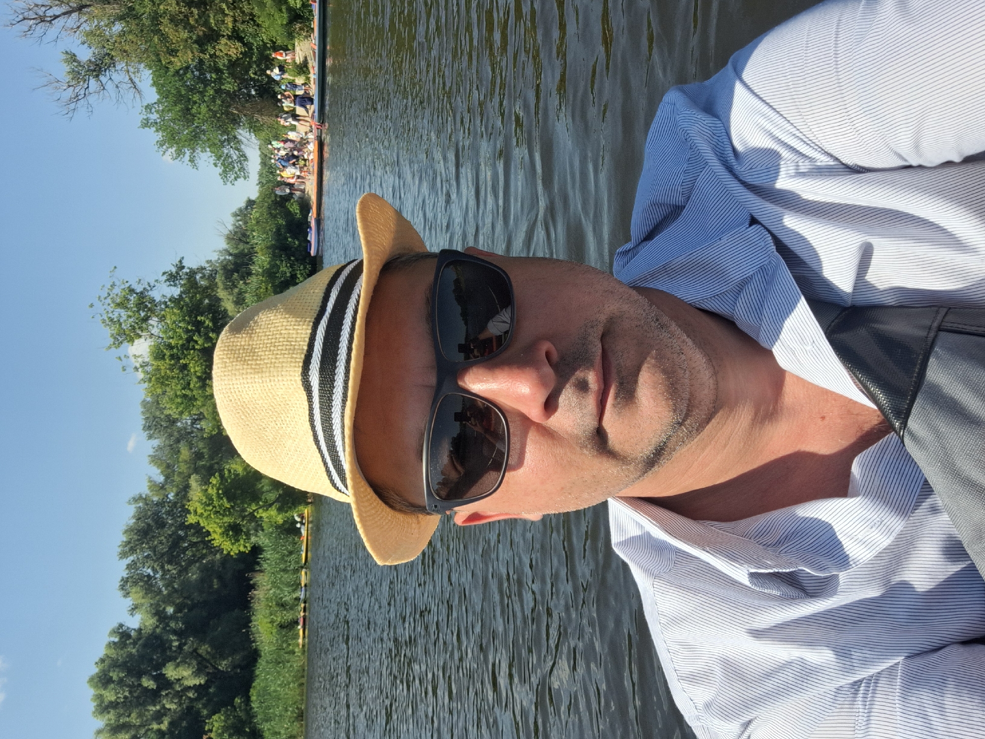 The image shows a man wearing a straw hat and sunglasses, taking a selfie on a body of water. In the background, there is a shoreline with trees and a group of people on what appears to be a beach, some of whom are on paddleboards or kayaks. The water is calm with ripples, and the sky is clear and blue. The man is wearing a light-colored, striped collared shirt. The location is identified as Potoky, Ukraine.