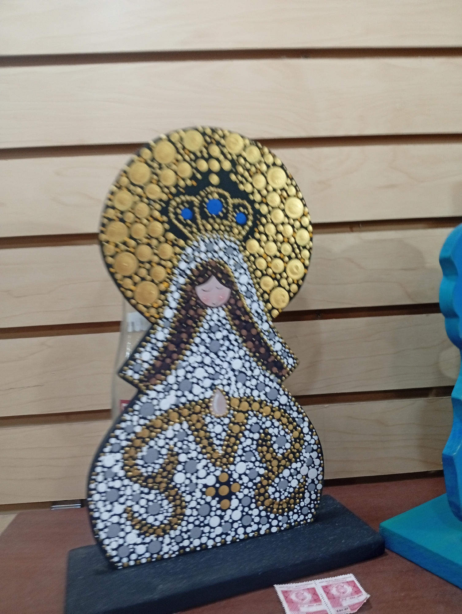 A decorative statue of a figure, possibly a religious icon, stands on a dark base. The figure is adorned with a large, circular halo decorated with gold and blue dots. The figure's face is simple, with closed eyes and rosy cheeks. Its body and head covering are intricately decorated with a pattern of white, grey, and gold dots. The statue is placed on a brown surface, with a light-colored wooden paneled wall in the background. To the right of the statue, a bright blue, abstract sculpture is partially visible. In the foreground, two small, pink stamps are lying on the brown surface. The overall scene suggests an indoor display, possibly in a shop or gallery.