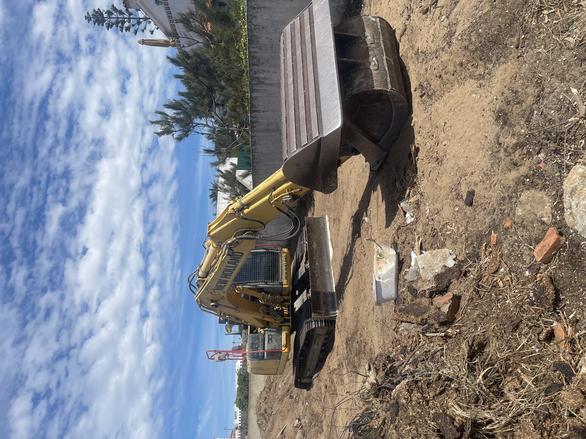 An excavator, a yellow New Holland model, is parked on a dirt lot in Colares, Portugal. The excavator is positioned to the left of the frame, with its bucket resting on the ground. In the background, a concrete wall runs horizontally across the image, with a white building and trees visible beyond it. The sky is blue with scattered white clouds. The ground is covered in sand and debris, including broken bricks and branches. A white tub is also visible in the foreground. The scene appears to be a construction or demolition site.