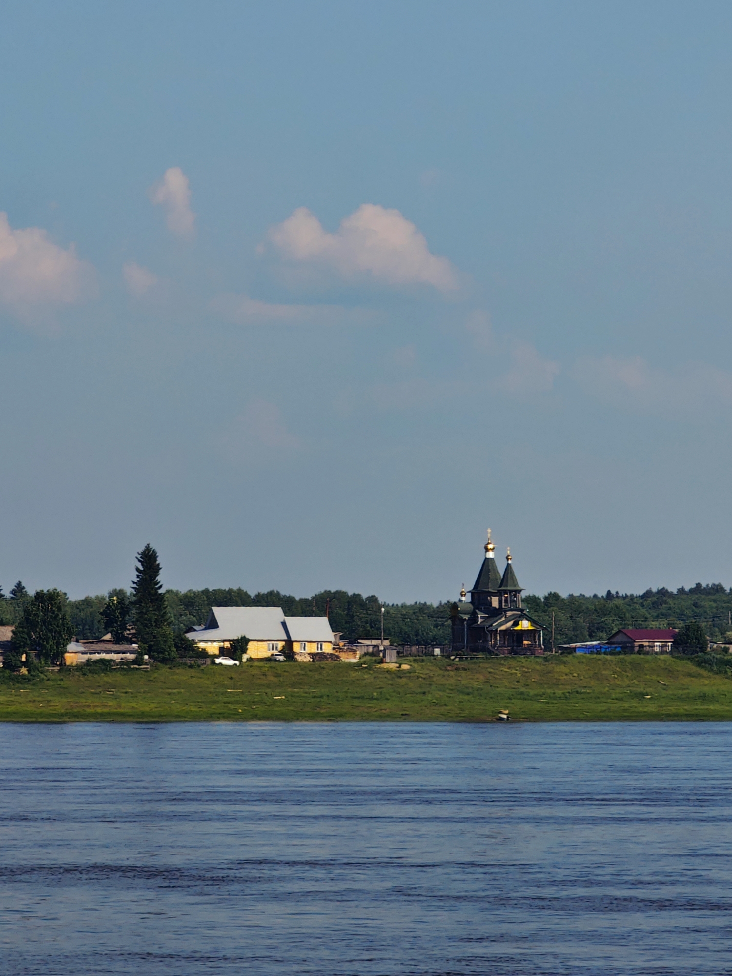 The image depicts a panoramic view of a village located on the banks of a wide river, likely in the city of Potapovo, Russia. The scene is dominated by a bright blue sky with scattered, fluffy white clouds. In the foreground, the choppy surface of a deep blue river fills the lower portion of the frame, reflecting the sky and suggesting a slight breeze.
The middle ground showcases a grassy embankment rising from the river. Several structures are visible on this elevated ground. To the left, a long, low yellow building with a light-colored roof is situated. A tall, dark green evergreen tree stands beside it, and a smaller evergreen is visible further left. A white car is parked in front of the yellow building.
Further to the right, a prominent dark wooden church with multiple golden domes stands out. The church features a complex architectural design with a steep roofline and intricate detailing. Adjacent to the church, a small building with a red roof and a bright blue structure are also present. A dense line of green trees forms a backdrop behind the village, extending across the horizon. The overall impression is of a serene, rural landscape on a clear day.