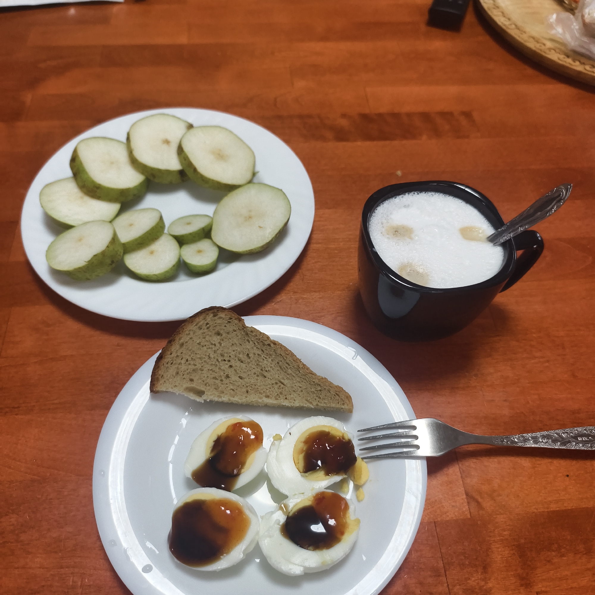 A close-up, high-angle image captures a meal arrangement on a reddish-brown wooden table in the city of Фоки, Russia. On the upper left, a white round plate contains approximately 13-15 slices of green-skinned fruit, appearing to be pears, cut into uniform rounds. In the bottom center, a second white round plate holds a single slice of dark, whole-grain bread positioned at the top. Below the bread, four halves of hard-boiled eggs are arranged, each with its yolk exposed and drizzled with a dark, viscous sauce; one egg half also features a reddish condiment. A silver fork with an ornate handle rests on the right edge of this plate, its tines near the eggs. To the right of the fruit plate and above the egg plate, a black ceramic mug contains a frothy, white beverage, likely a latte or cappuccino, with light brown spots on its surface. A silver spoon with a decorative handle is submerged in the drink. In the upper right background, a dark, rectangular remote control is partially visible next to a section of a round wooden tray adorned with carved patterns, which contains an object encased in clear plastic. No people are present in the scene.