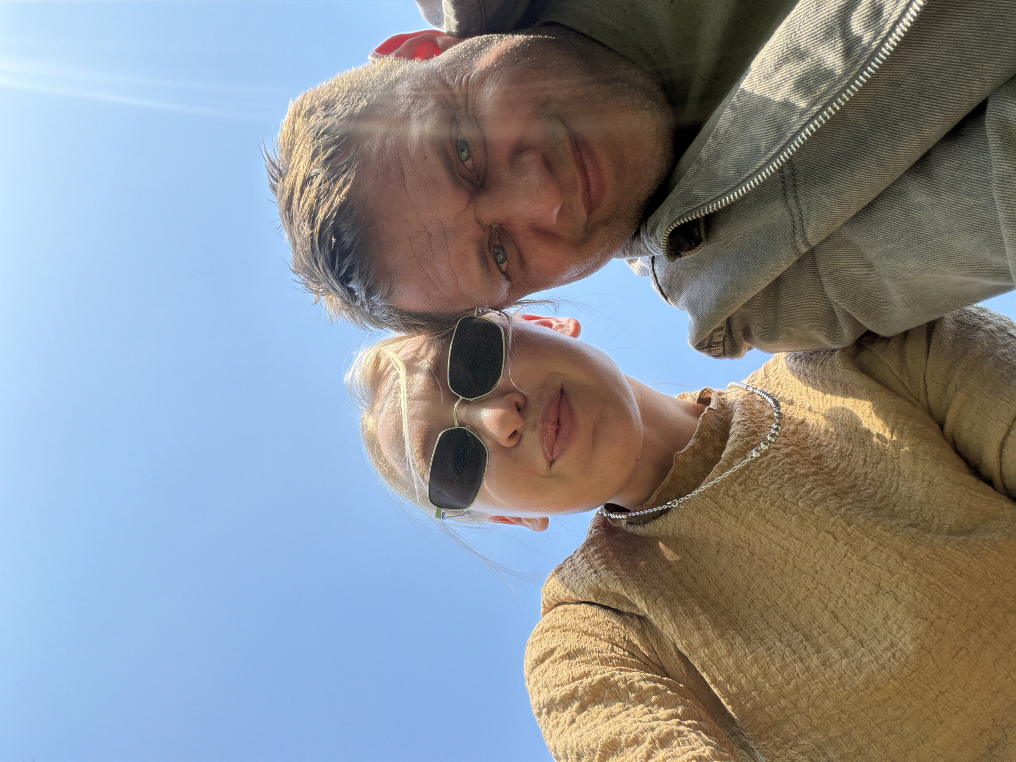 A low-angle shot captures a man and a woman posing for a photo against a clear blue sky. The man, positioned at the top of the frame, has short, light brown hair, visible wrinkles, and is wearing a zipped-up, light green jacket. The woman is wearing sunglasses and a gold-colored top with a silver chain necklace.
There is no visible information to identify the location as Chaikovsky, Russia.