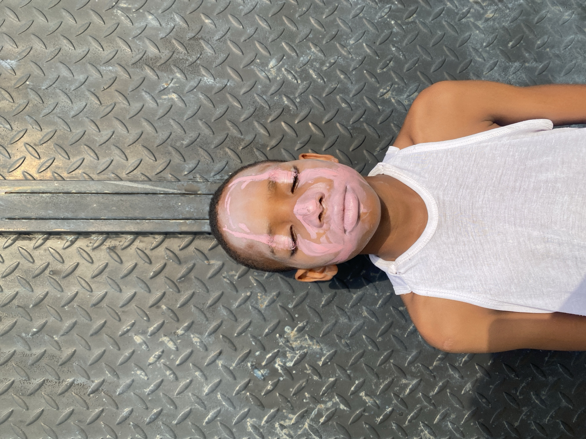 A child lies on a grey metal surface patterned with raised, elongated ovals. The child wears a white tank top and has pink cream spread over their face. The child is lying on their back with their head turned towards the viewer's left. The location is unknown, and there is no evidence to suggest that this image was taken in Iranla, Nigeria.