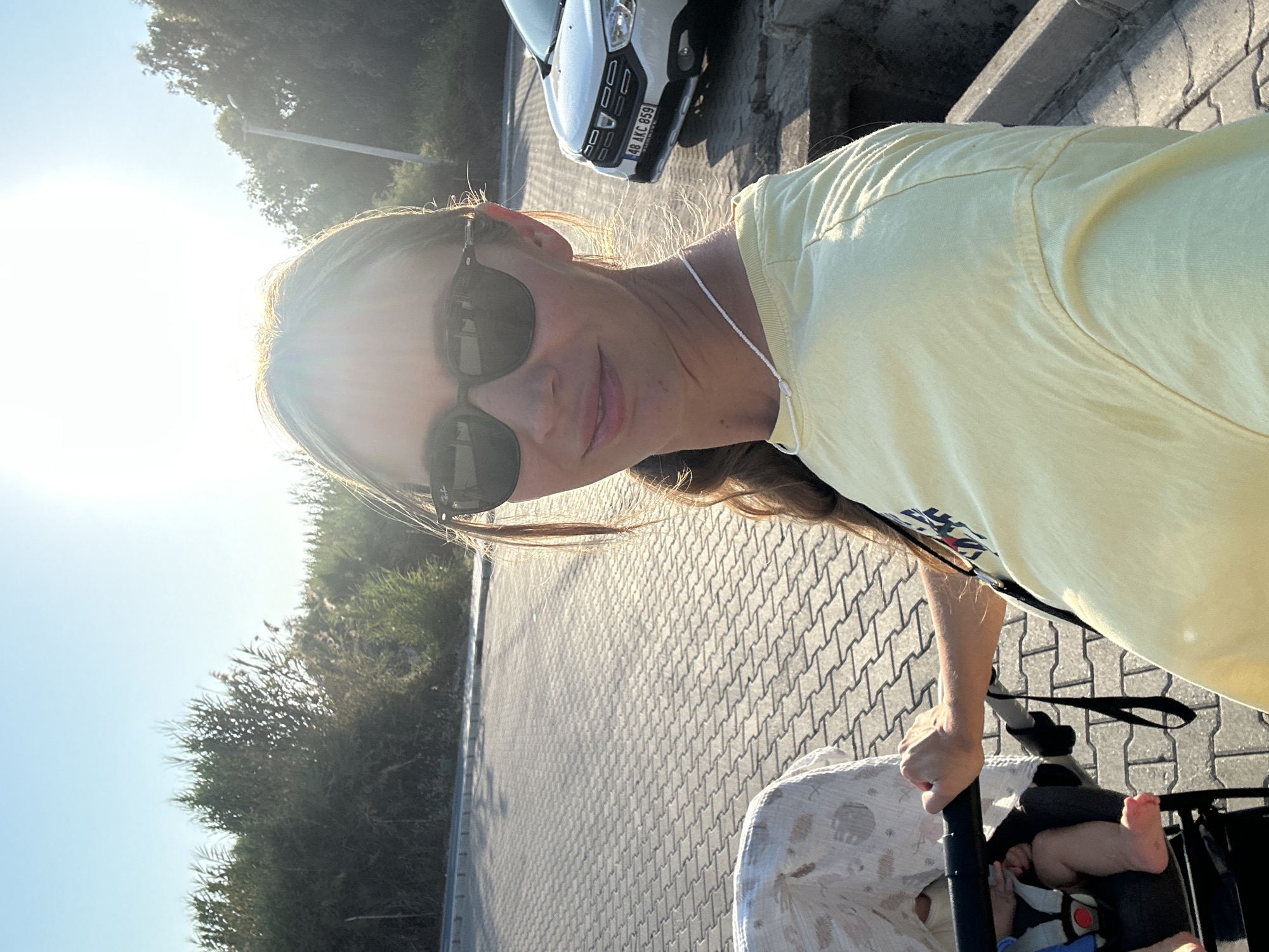 A woman wearing sunglasses and a light yellow t-shirt takes a selfie while holding the handle of a stroller. A baby is visible in the stroller, wearing a blue and grey harness. A white car with a Turkish license plate (48 AKC 859) is parked to the right of the woman. The scene appears to be outdoors on a paved area, possibly a walkway or parking lot, with trees and vegetation in the background. The sun is bright and positioned high in the sky, casting shadows. The presence of the Turkish license plate suggests the location is in Turkey, and given the sunny weather and general appearance, it could be a coastal area like Fethiye.