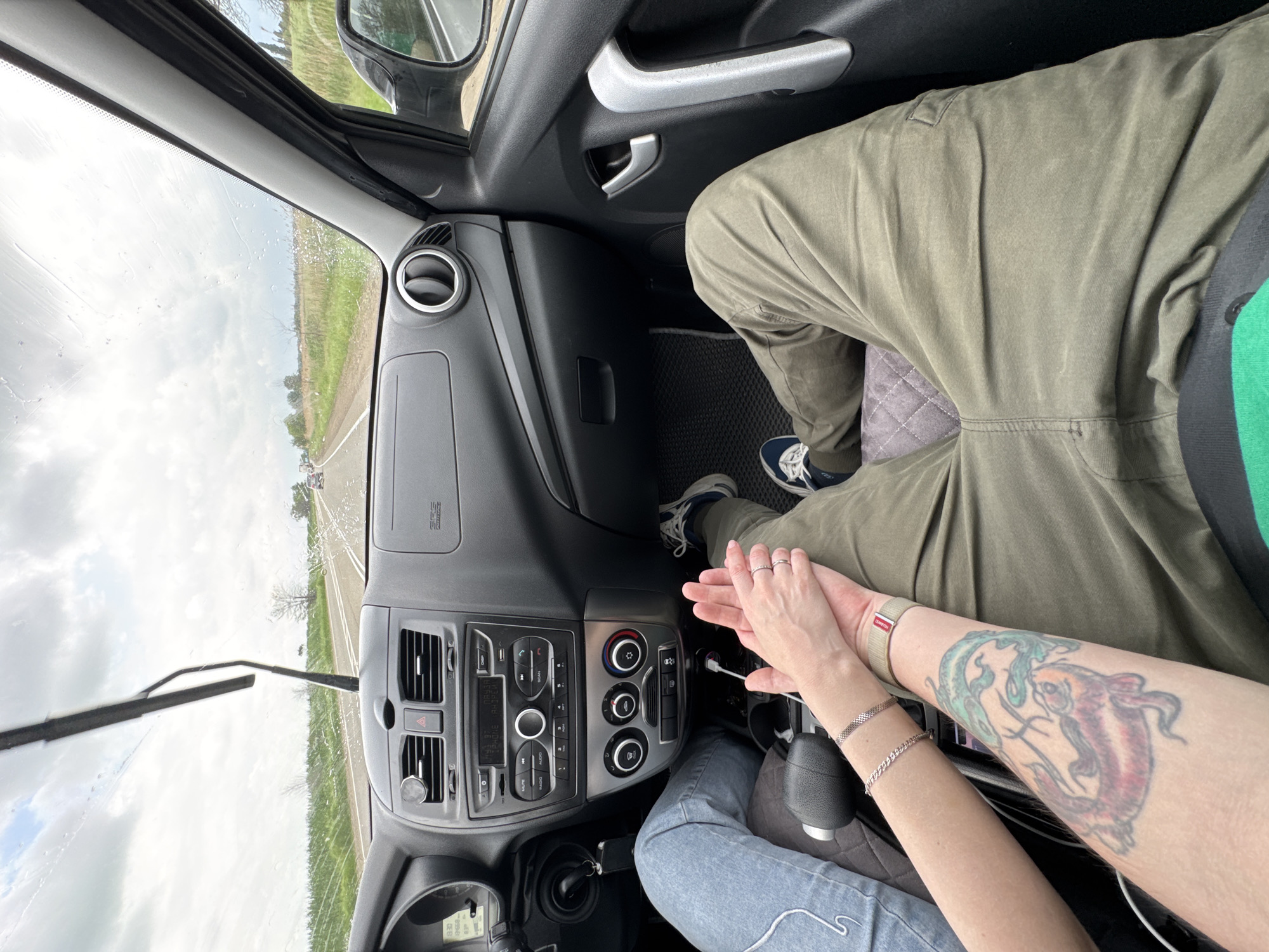 The image shows an interior view of a car, likely in motion, with a view of the road and fields through the windshield. The location is identified as Novomyshastovskaya, Russia. The windshield wipers are active, indicating rain. The dashboard and center console of the car are visible.
Two people are present. One person is wearing olive green pants and blue and white sneakers. The other is wearing blue jeans. Their hands are clasped together. The person wearing jeans has a tattoo of koi fish on their forearm, and is wearing a bracelet.