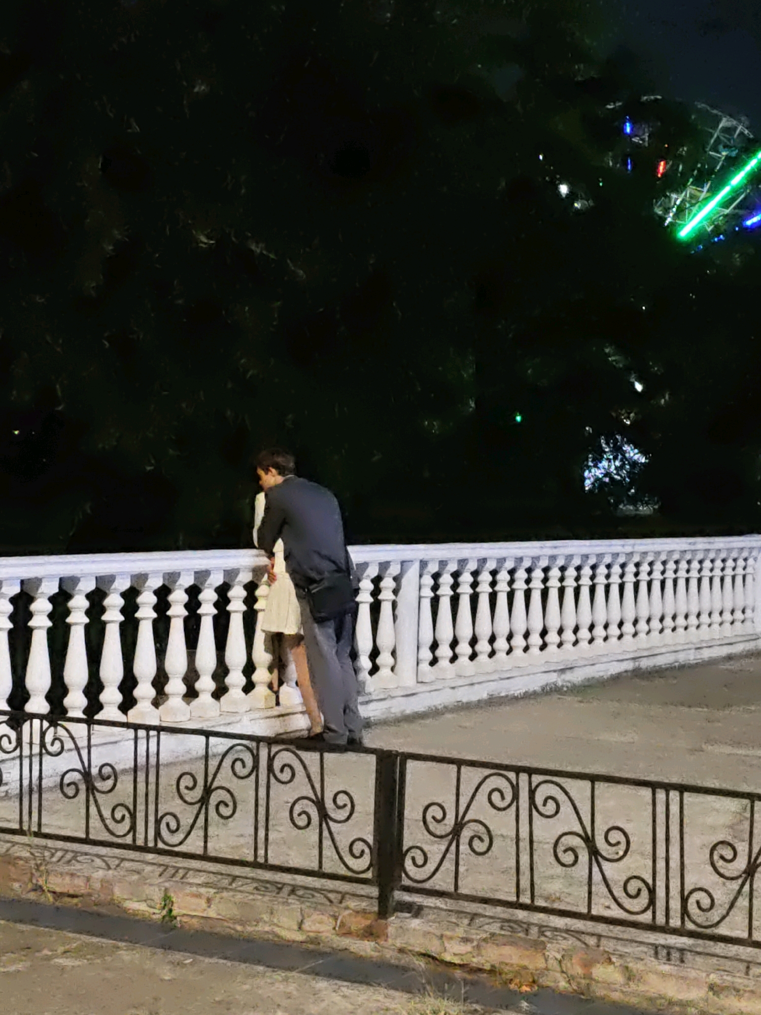 The image shows a couple standing by a white ornate railing at night. The man is wearing a dark jacket and gray pants, and the woman is wearing a light-colored dress. They appear to be facing away from the camera, looking out at something in the distance. In the background, there are trees and the lights of a Ferris wheel, suggesting an amusement park or entertainment venue. The scene is illuminated by artificial lights, creating a somewhat dark and moody atmosphere. The white railing and the black wrought-iron fence in the foreground are prominent elements of the scene. Based on the context of the city of Rostov-on-Don, Russia, this scene likely depicts a romantic moment on an evening stroll near a park or amusement area.