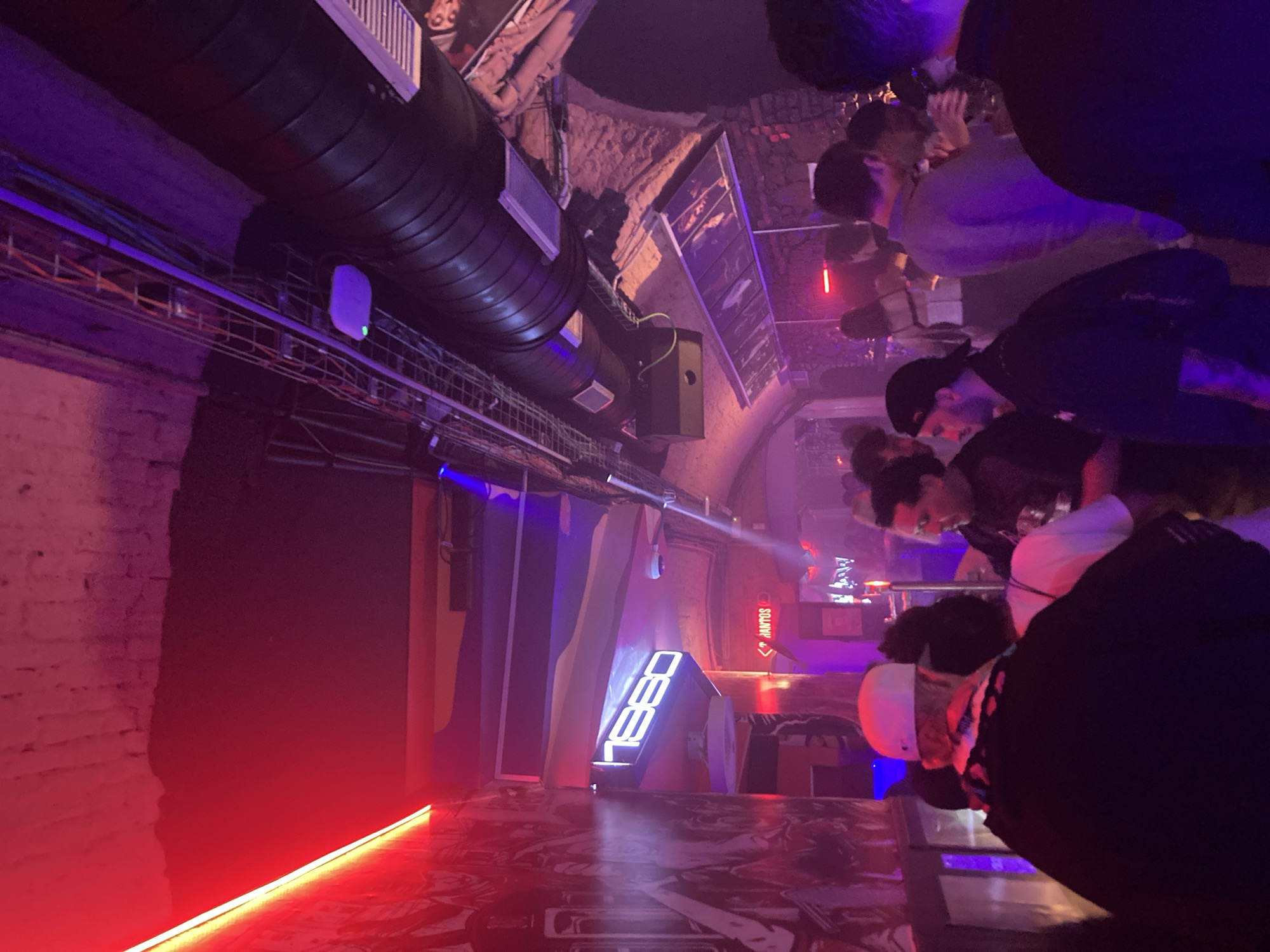 The image captures the interior of Heso Barcelona, a club located in Barcelona, Spain. The environment is characterized by an exposed brick ceiling with arched architectural features and an industrial aesthetic. Overhead, dark, cylindrical ventilation ducts run across the ceiling, accompanied by metal trussing holding an array of lighting fixtures, speakers, and exposed cabling.
The illumination is predominantly atmospheric, featuring a prominent red neon strip light along the upper left wall, casting a deep red glow on a large, abstract mural below it. Additional purple and blue ambient lighting emanates from various sources, including spotlights and ceiling-mounted fixtures, creating a dynamic light environment. A prominent, illuminated sign displaying "HESO" in a digital segment-style font is mounted on a structural column or wall section on the left side of the frame. Further back, a partially visible red illuminated sign reads "MANTOS".
Numerous individuals are present, standing in groups and conversing throughout the space. In the immediate foreground, the back of a person wearing a white baseball cap and a dark jacket is visible. To their right, another person in a white t-shirt is partially seen. In the midground, a man with a dark beard and a dark shirt holds a drink and engages with another individual. Next to him, a man wearing a black baseball cap is seen in profile. Various other patrons are dispersed across the midground and background, most holding drinks. Their interactions appear social, consistent with a nightclub or bar setting.