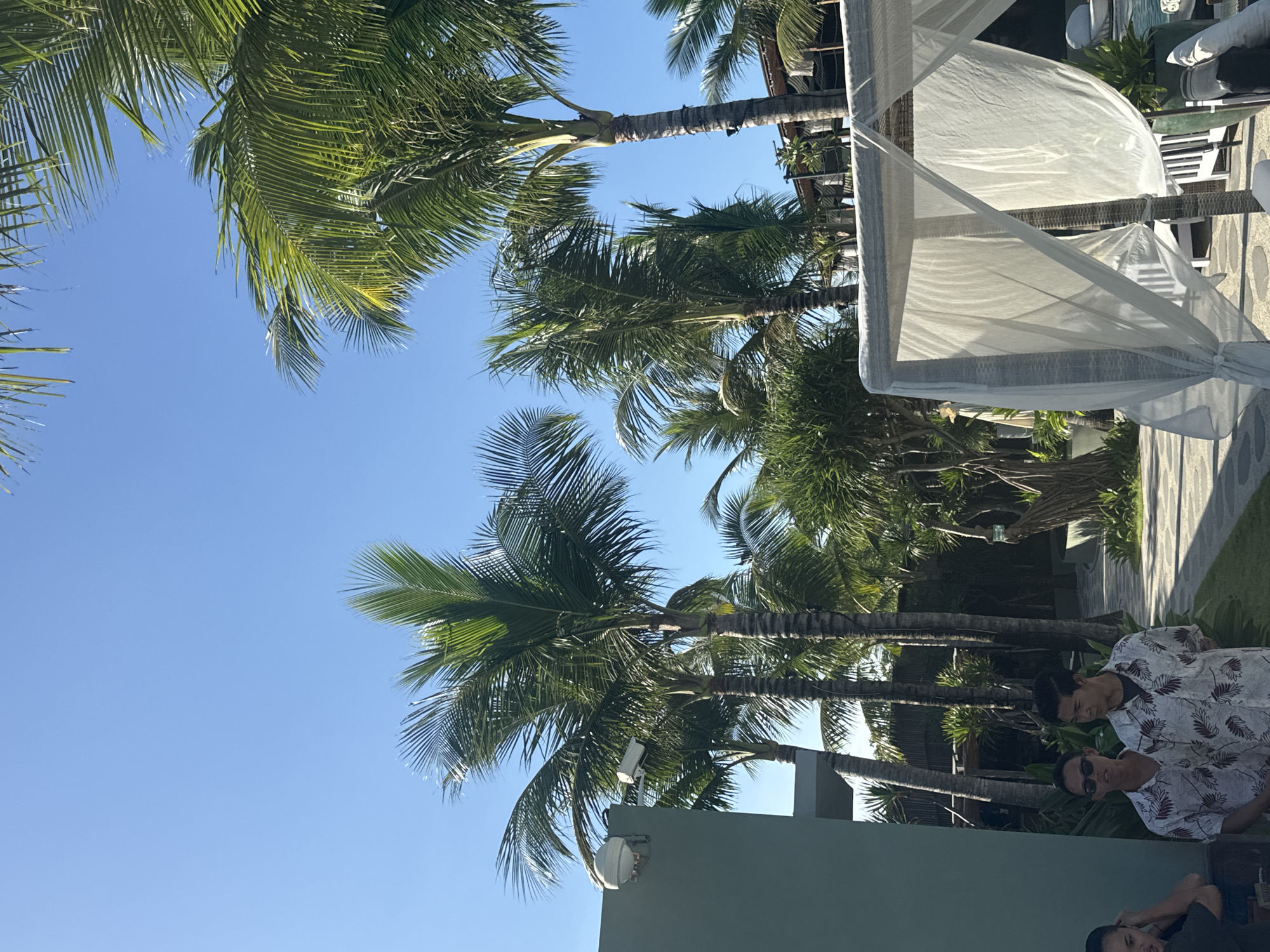 The image captures a scene in Sukawati, Indonesia, under a bright blue sky. Palm trees are visible, stretching upwards. A white canopy structure occupies the right side of the image. Two men are seen near the bottom of the frame, one wearing sunglasses and a patterned shirt. A building is partially visible at the bottom edge of the photo.