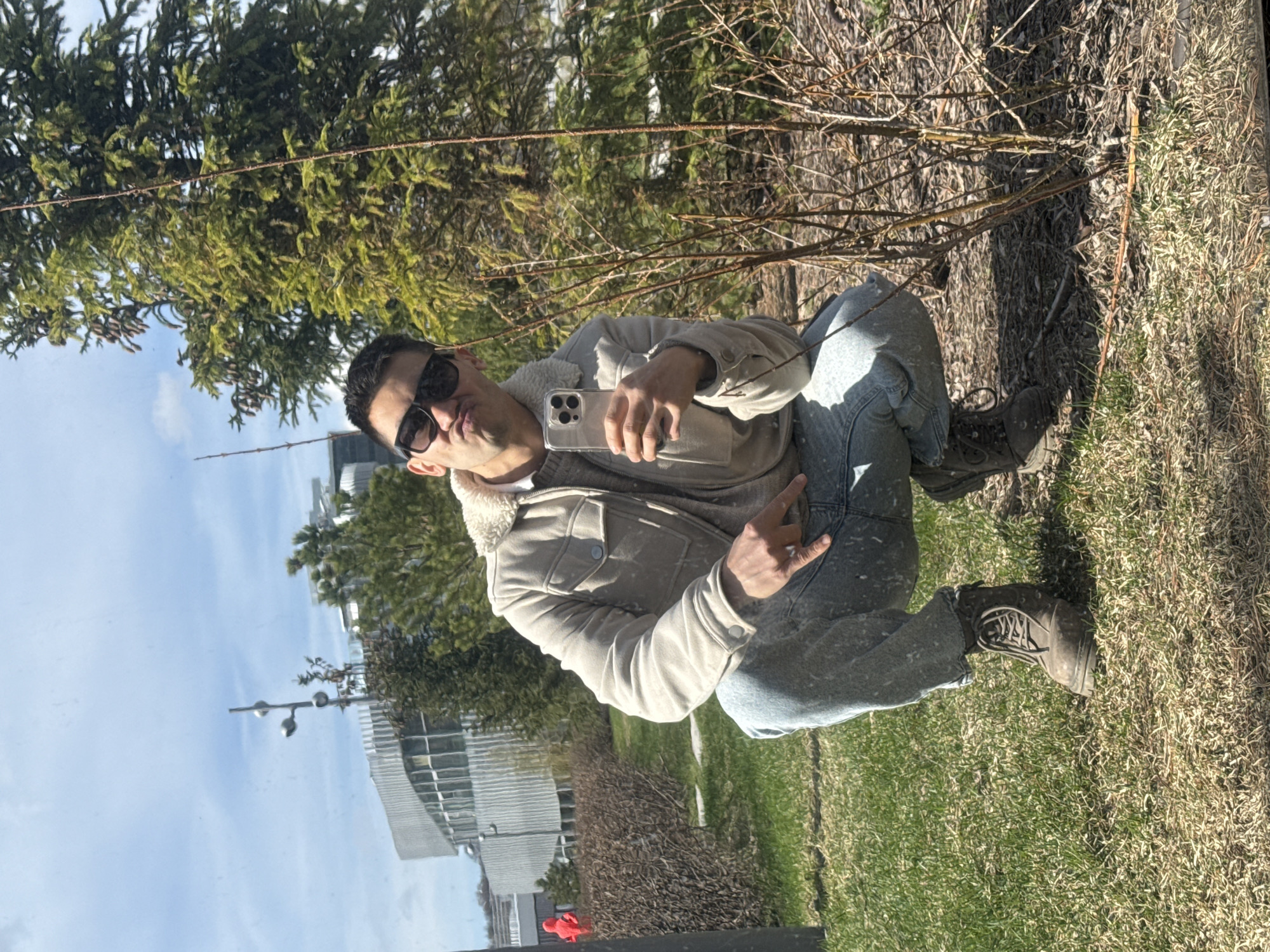 A man is squatting in a park, taking a selfie with his smartphone. He is wearing a light-colored jacket with a furry collar, jeans, and boots. He is also wearing sunglasses. Behind him are trees, bushes, grass, and a building that suggests he is in a city. Considering the request to locate the image in the city of Kazan, Russia, and the visual characteristics of the buildings, this could possibly be near a building with modern architecture in a park setting, consistent with urban landscapes in Kazan. The photo appears to be taken in natural sunlight, possibly on a clear day. The man is holding his phone in front of him, posing for the photo. The buildings visible in the background suggest a mix of modern architectural styles, commonly found in urban settings like Kazan.