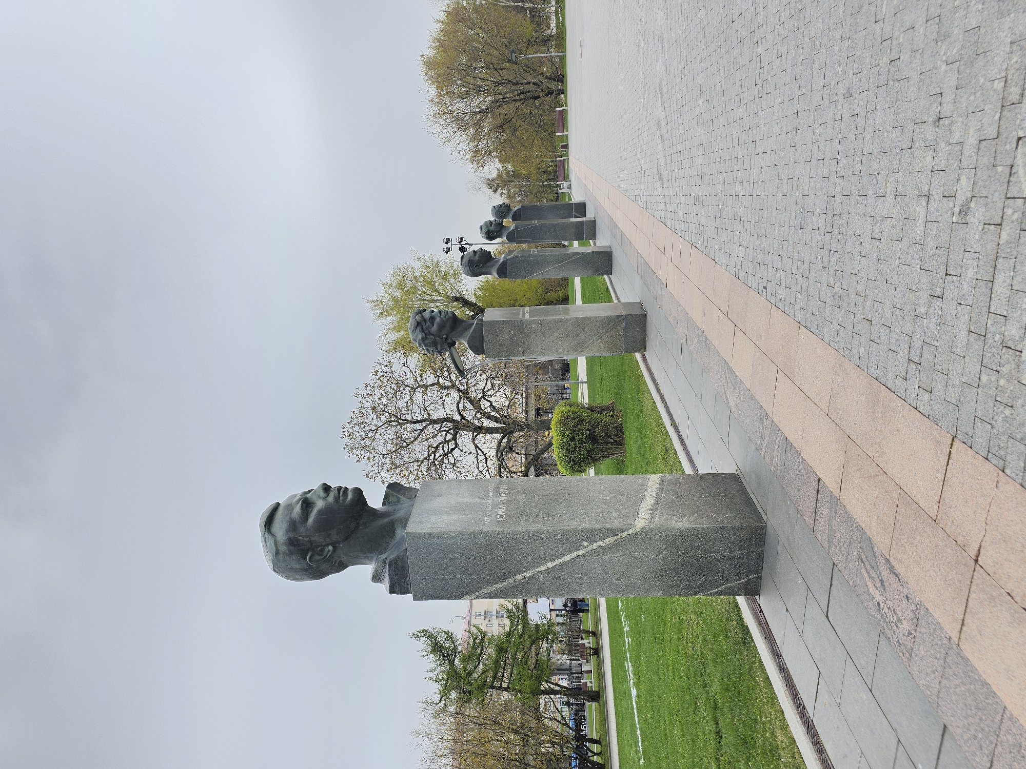 The image displays a row of bust statues positioned along a paved pathway, likely in a park or memorial area. In the foreground, the most prominent statue features a dark, possibly bronze, bust of a male figure looking upwards. This bust is mounted on a tall, rectangular pedestal made of dark grey mottled granite. Inscribed on the pedestal in Cyrillic are the words: "ЛЕТЧИК-КОСМОНАВТ СССР" (Pilot-Cosmonaut of the USSR) and "ЮРИЙ ГАГАРИН" (Yuri Gagarin), identifying the subject as the Soviet cosmonaut Yuri Gagarin.
Behind the first bust, several other similar busts on pedestals recede into the distance along the pathway, suggesting a commemorative alley. The pathway itself is paved with light grey interlocking pavers, bordered by a strip of reddish-brown stone. To the left of the busts, there is a wide area of green grass and several deciduous trees, some with new, light green leaves, indicating springtime. In the background, beyond the trees, blurry urban buildings are visible. The sky is overcast and grey, providing diffused daylight. The scene depicts a static memorial, with no specific activity taking place, conveying a sense of solemnity and remembrance in Moscow, Russia.