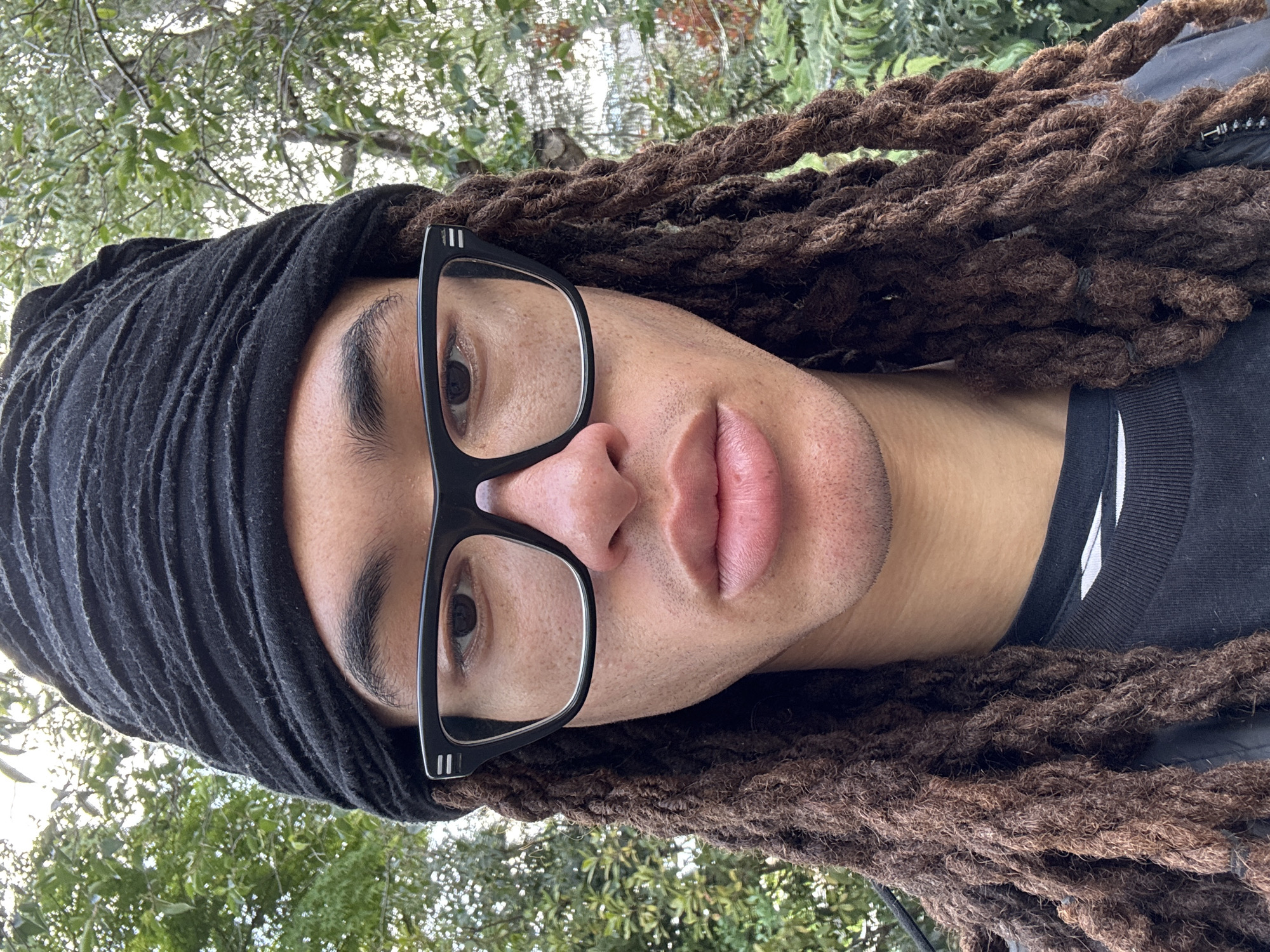 A close-up portrait shows a person with brown dreadlocks and a black head covering. They wear black-framed glasses. The person is outdoors, and blurred foliage is visible in the background. The image was likely taken in the city of Les Ulis, France, but no specific landmarks or elements confirm this.