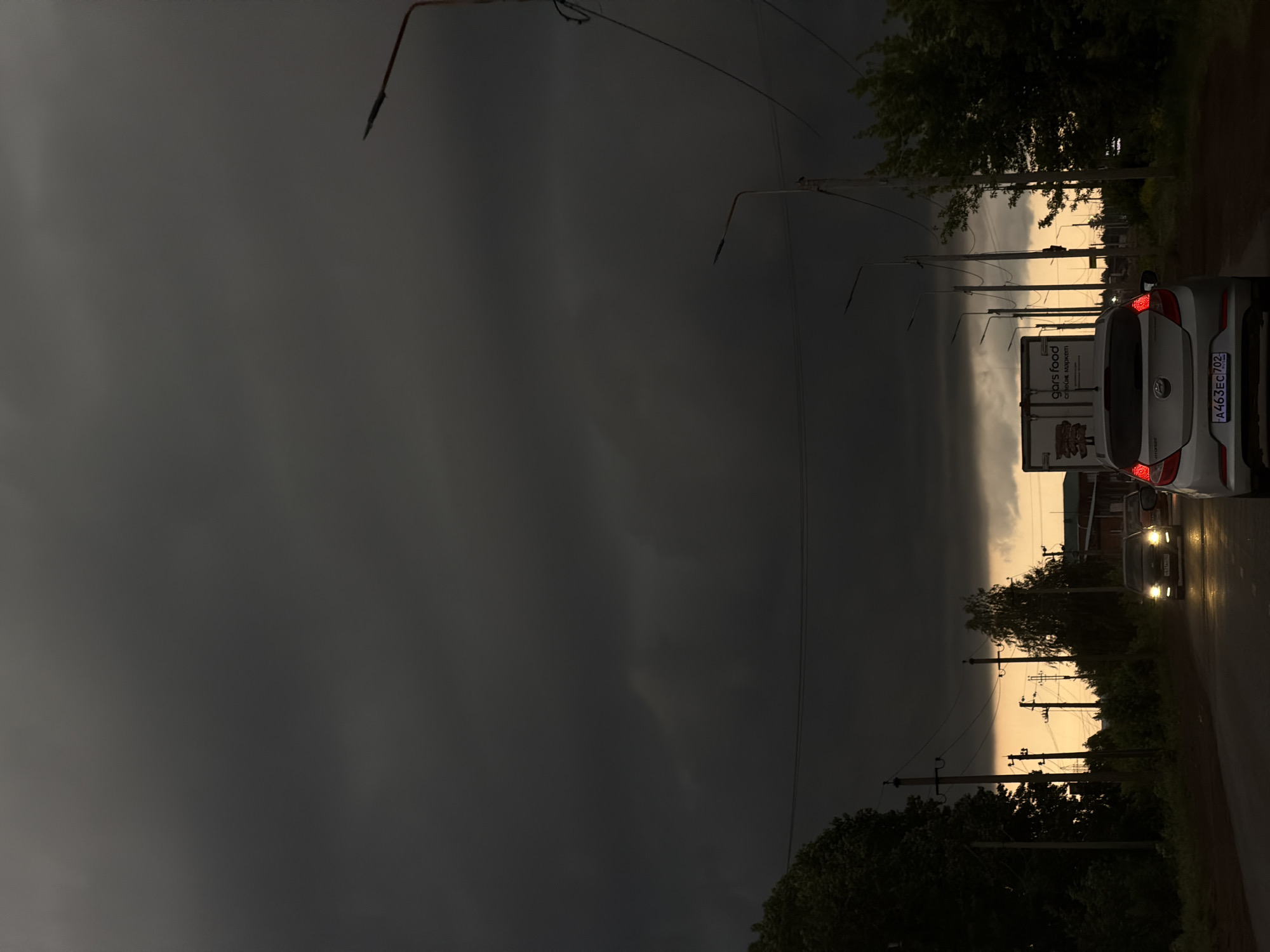 A photograph shows a street scene in Neftekamsk, Russia, dominated by a looming dark storm cloud in the sky. A white van is parked on the right side of the road, displaying the license plate "A463EC702" and a "Gorsfood" advertisement on its rear. A car with illuminated headlights approaches from the opposite direction. Utility poles and trees line the road, silhouetted against the brighter sky not yet obscured by the storm.