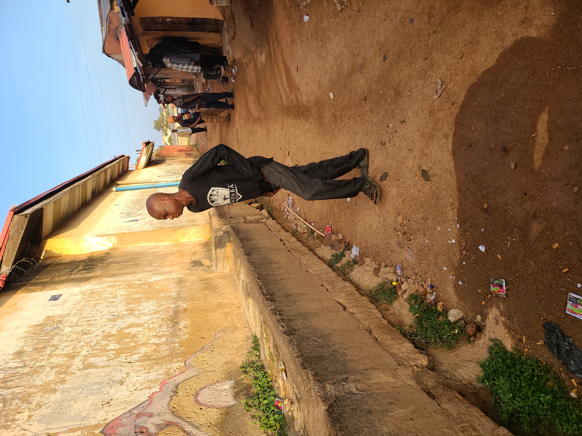 A man wearing a black long-sleeved shirt and dark pants balances in a plank position on a dirt path alongside a low concrete wall and weathered building in Jos, Nigeria. A group of people stand in the background near a row of buildings under a partly sunny sky. Debris litters the dirt path and the area between the path and the wall.